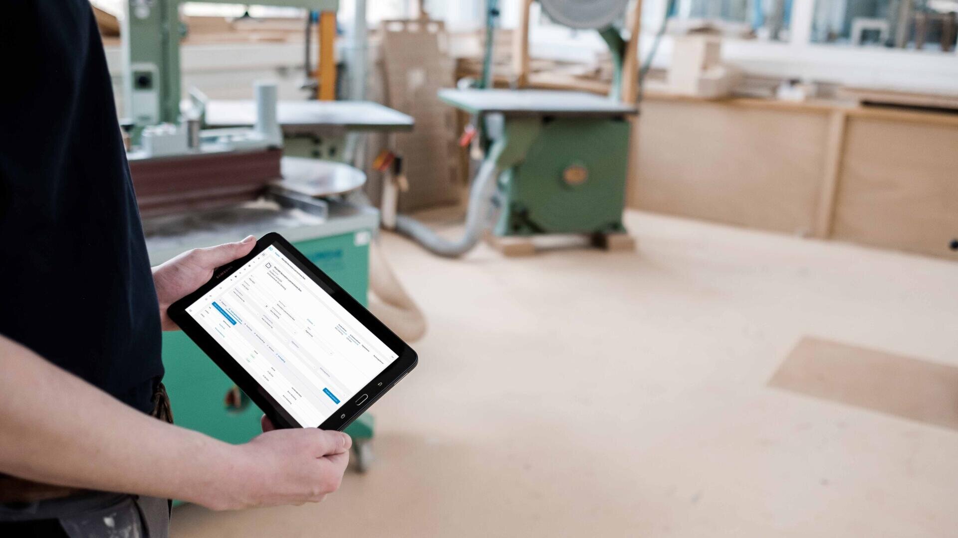 Eine Person hält ein Tablet mit einer digitalen Benutzeroberfläche in einer Werkstatt mit grünen Industriemaschinen und Holzoberflächen im Hintergrund.