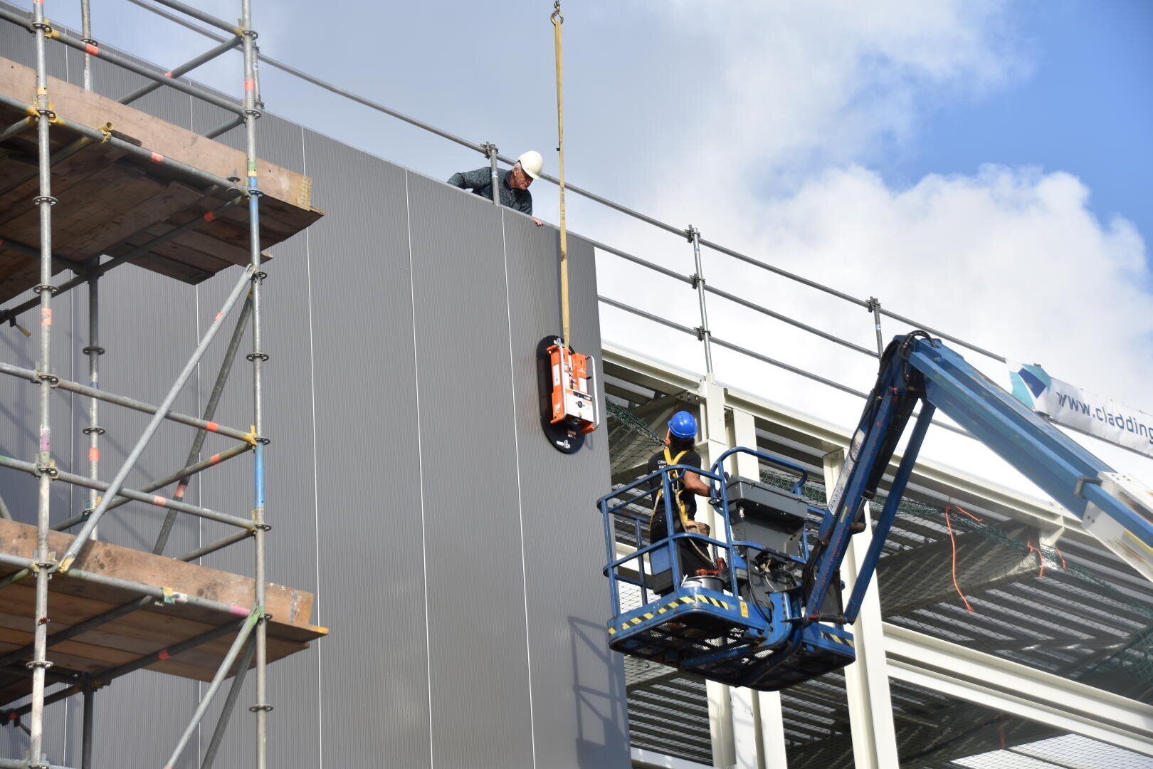 Deux ouvriers du bâtiment installent un grand panneau gris sur l'extérieur d'un bâtiment à l'aide d'une grue et d'une plate-forme de travail surélevée bleue, avec des échafaudages et des structures métalliques visibles autour d'eux.