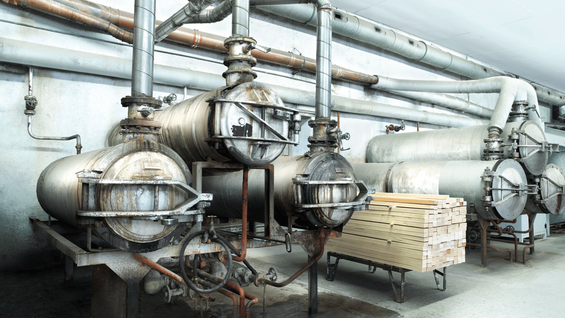 Reihen von industriellen Metalldruckbehältern und -rohren in einer Fabrikumgebung, mit einem Stapel zugeschnittener Holzbretter in der Nähe auf dem Boden. Der Bereich wirkt sauber, aber industriell und utilitaristisch.