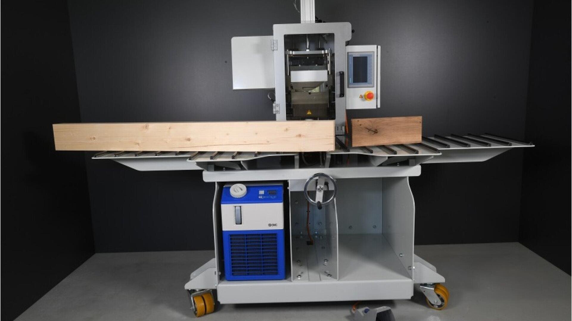 Une machine à travailler le bois avec une planche de bois clair sur ses rouleaux de transport, avec un panneau de commande numérique, des boutons et des roues, sur un fond sombre.