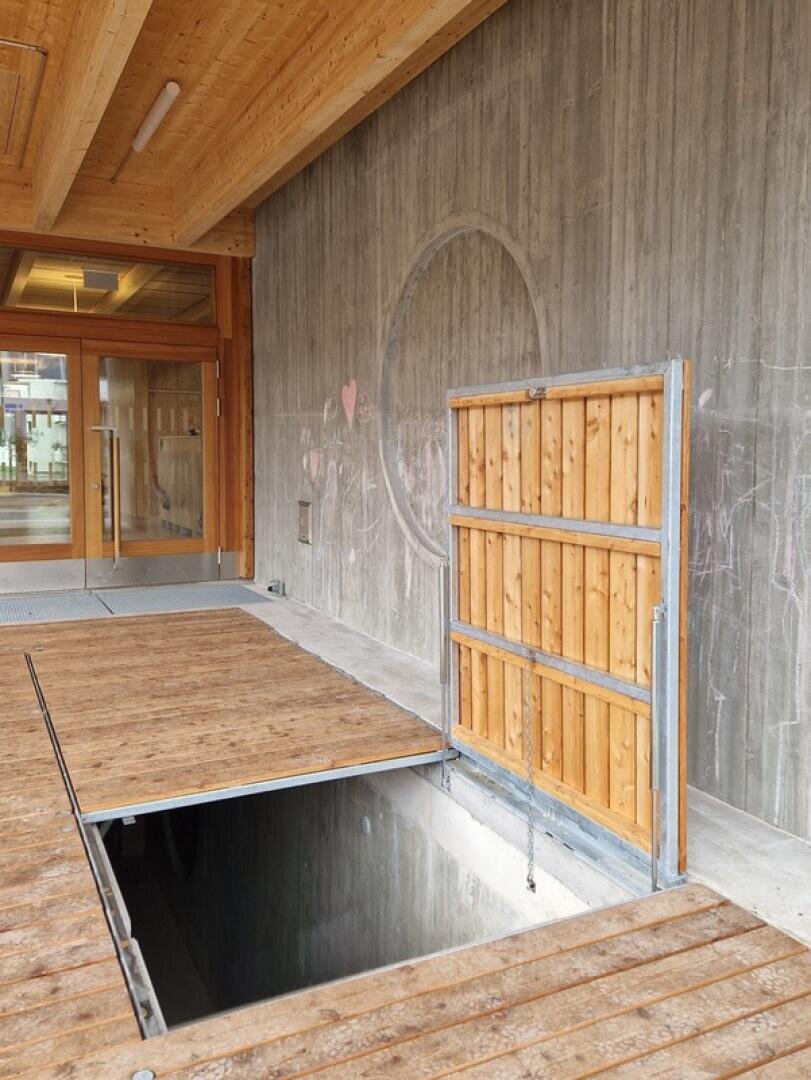 Eine hölzerne Plattform neben einer Wand ist mit einem Scharnier geöffnet, das ein Loch zu einem unterirdischen Bereich freigibt. Die Wand ist aus Beton mit hellen Kreidezeichnungen, und im Hintergrund befinden sich große Glastüren.