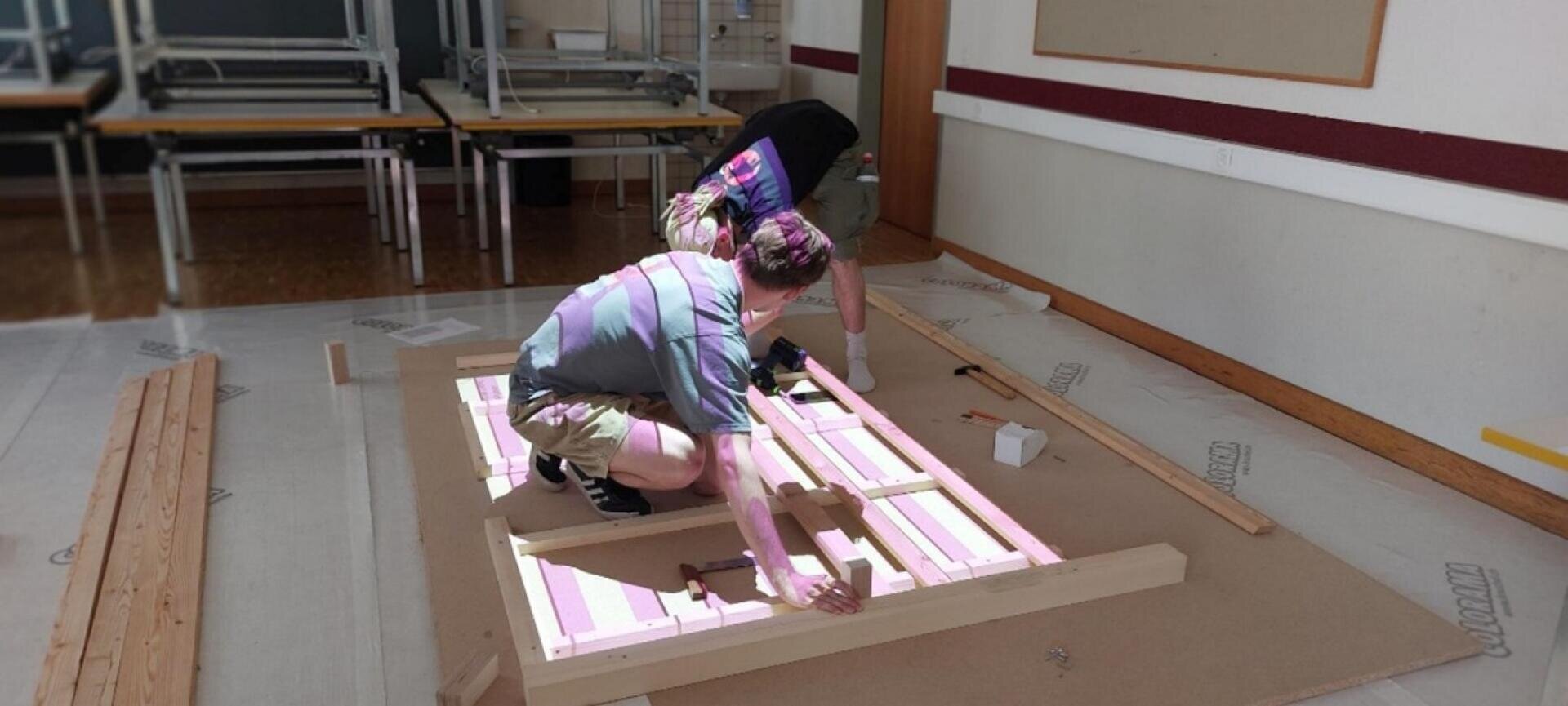 Dans une salle de classe, deux personnes assemblent un cadre en bois sur le sol, entouré de planches de bois et de tables empilées au fond. Une personne s'agenouille pendant que l'autre regarde ou assiste.