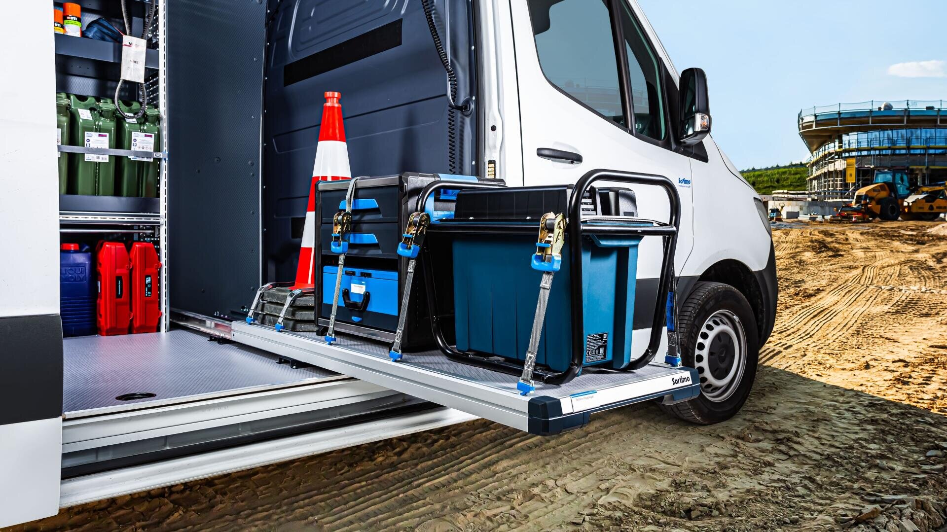 Ein weißer Lieferwagen mit geöffneter Seitentür zeigt organisierte Ausrüstung, darunter blaue Lagerbehälter, die auf einem Schiebetablett befestigt sind, und einen orangefarbenen Verkehrskegel, der auf einer sandigen Baustelle mit Gebäuden im Hintergrund geparkt ist.