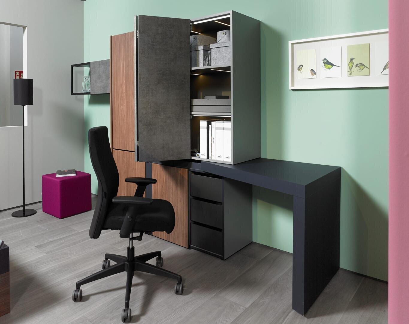 Bureau à domicile moderne avec une chaise pivotante noire, un bureau minimaliste et des armoires. Des étagères à l'intérieur d'une armoire ouverte contiennent des boîtes et des dossiers. La pièce a des murs vert menthe, un sol en bois et un tableau d'oiseaux encadré au mur.