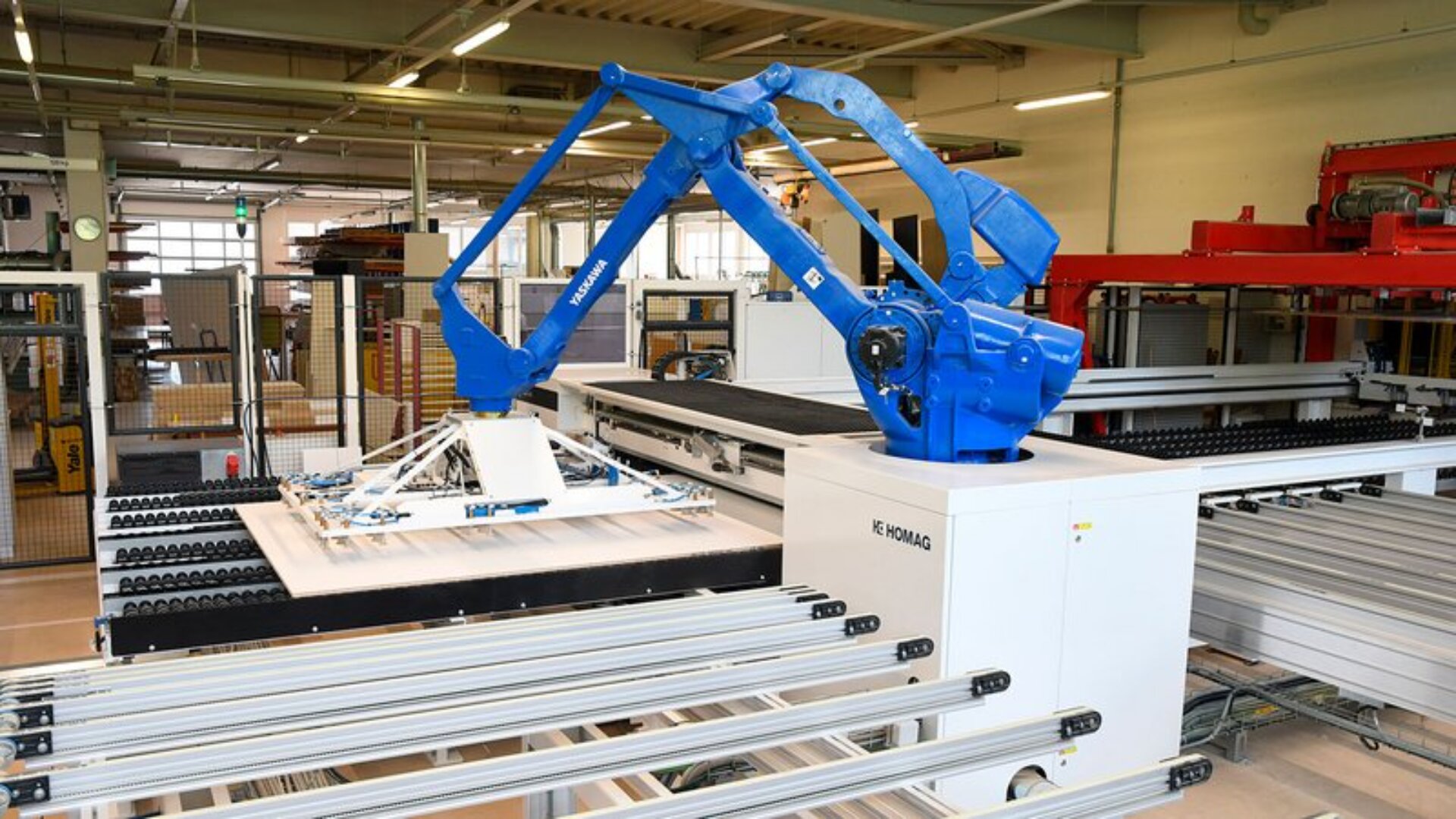 Ein blauer Industrieroboterarm arbeitet in einer modernen Fabrik und bearbeitet flache Platten oder Bretter auf einer weißen Maschine, die von Metallschienen und Fördersystemen umgeben ist. Die Umgebung ist sauber und gut beleuchtet.
