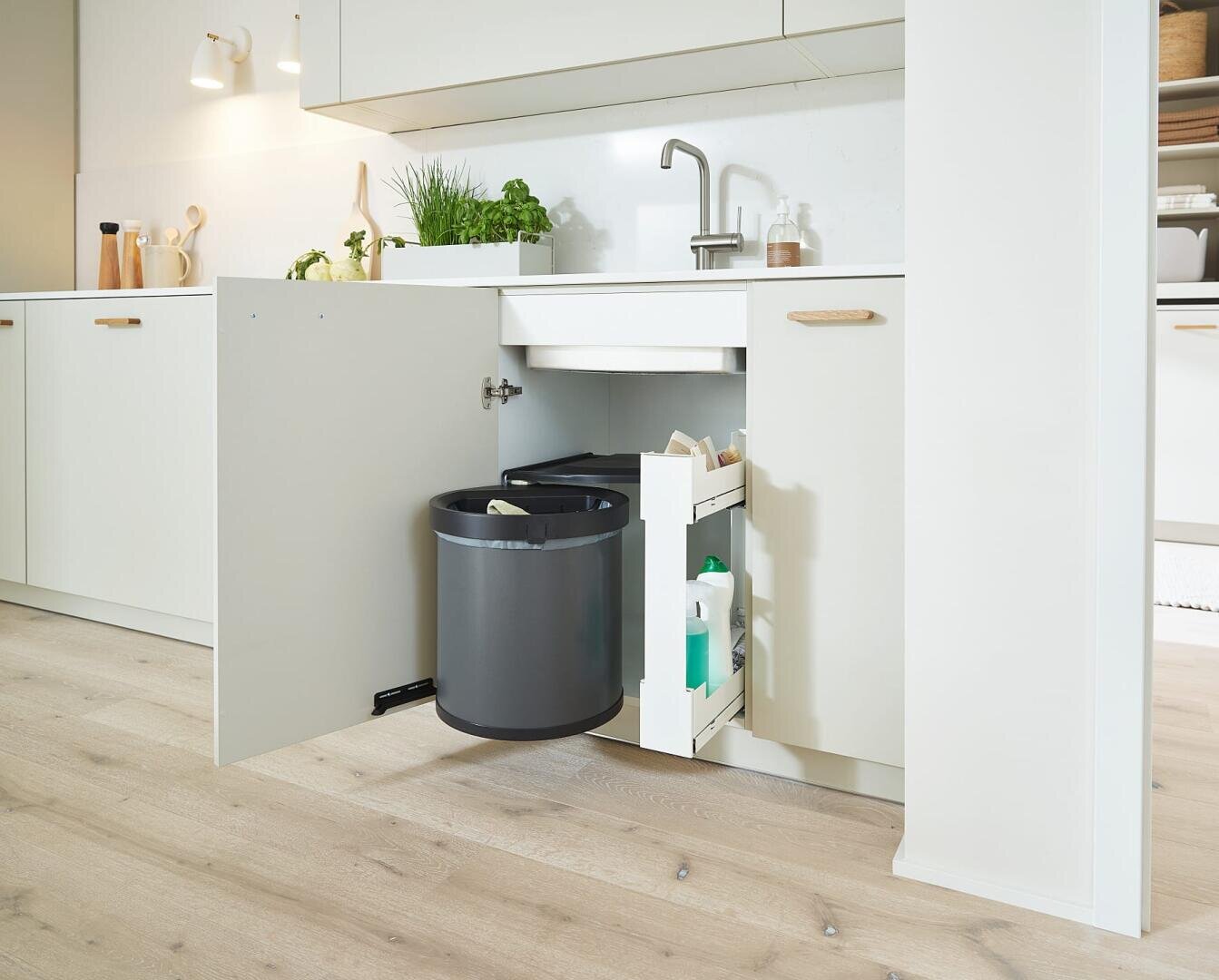Eine moderne Küche mit einem ausziehbaren Schrank unter der Spüle, in dem sich ein runder Mülleimer und Stauraum für Reinigungsmittel befindet, vor hellen Schränken und einem Holzboden.