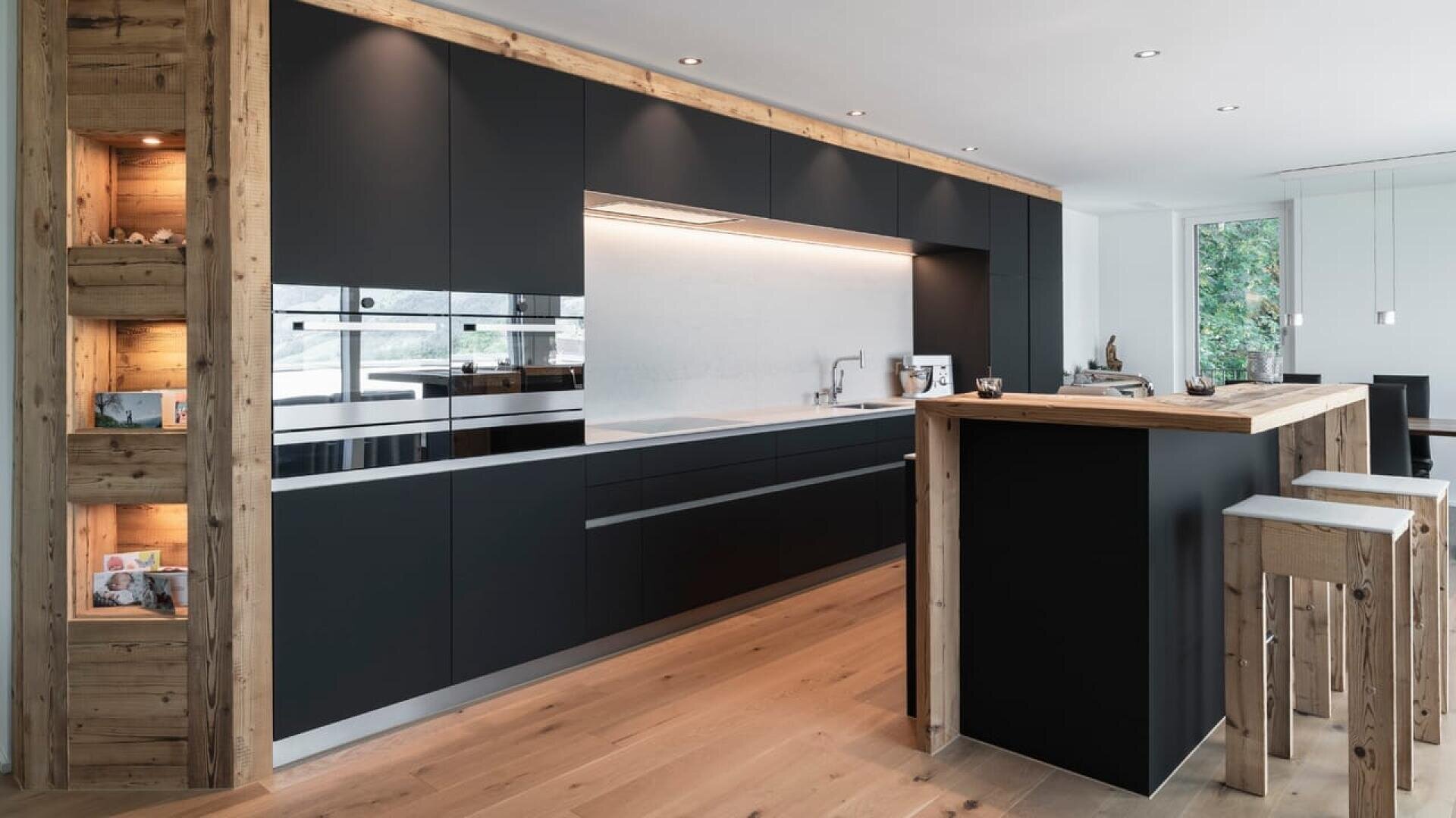 Cuisine moderne avec des armoires noires mates, des appareils encastrés et des touches de bois clair. Un îlot en bois avec des tabourets assortis et des étagères ouvertes ajoute de la chaleur. De grandes fenêtres apportent de la lumière naturelle.