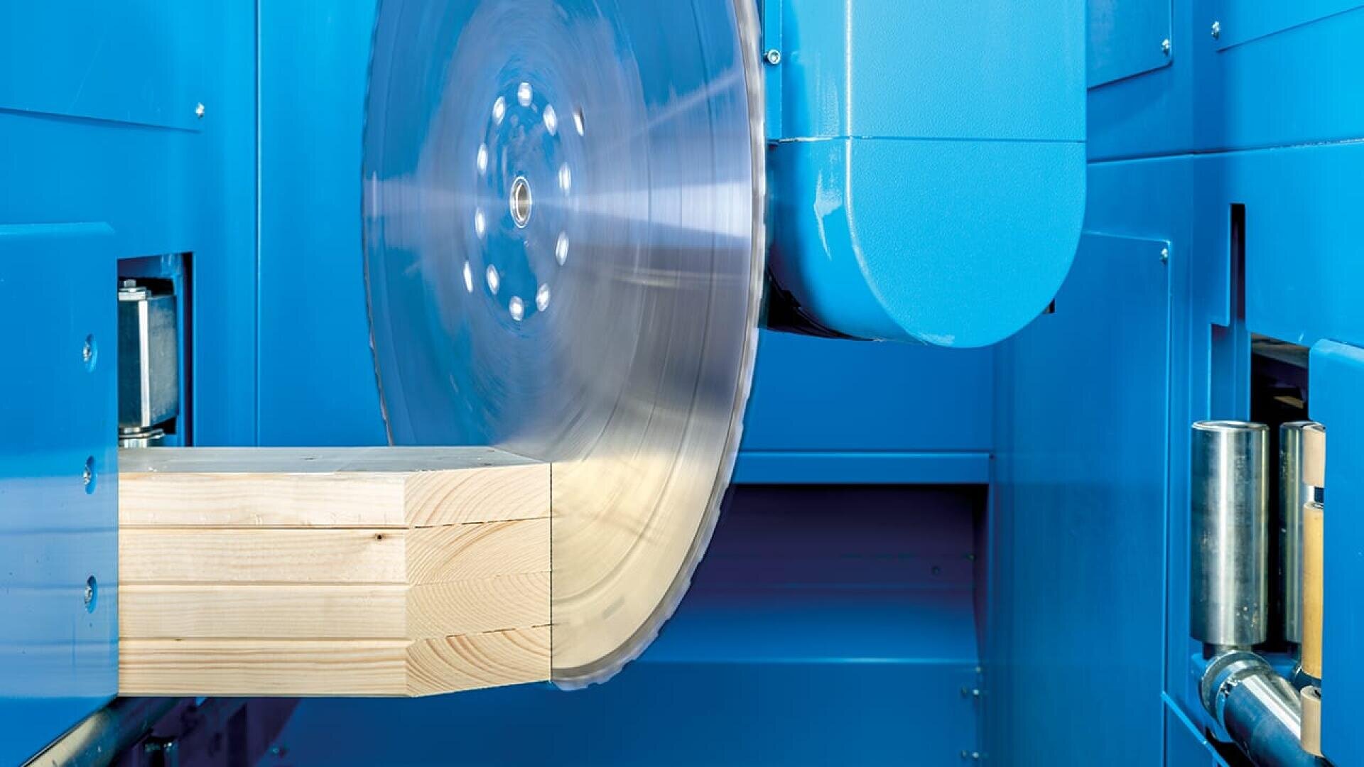 Ein großes Kreissägeblatt in einer blauen Industriemaschine schneidet durch einen dicken Holzbalken.