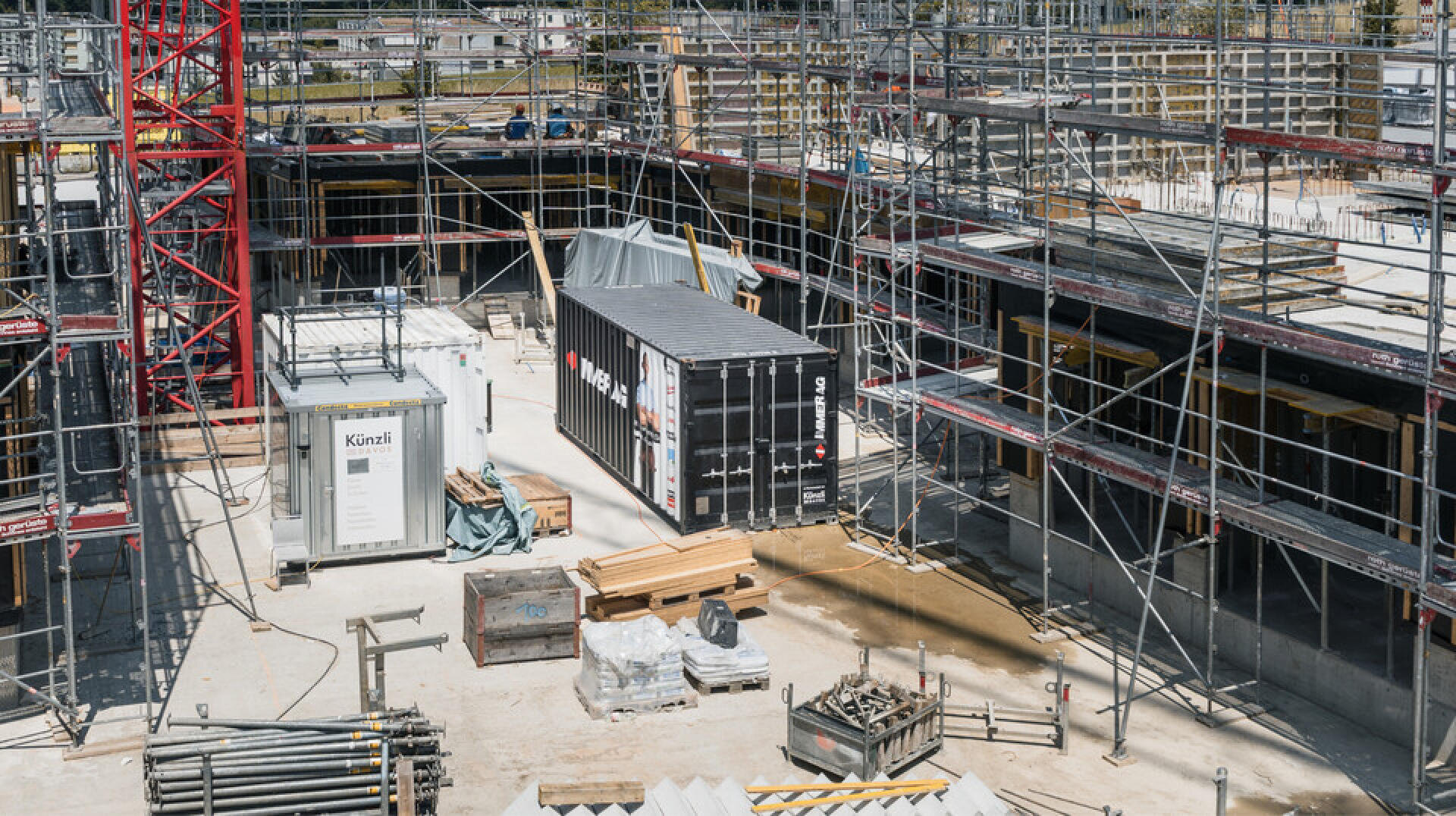 Baustelle mit Gerüsten, einem roten Kran, Frachtcontainern, Materialstapeln und verstreut auf einem Betonfundament stehenden Geräten. In der Szene sind keine Arbeiter zu sehen.