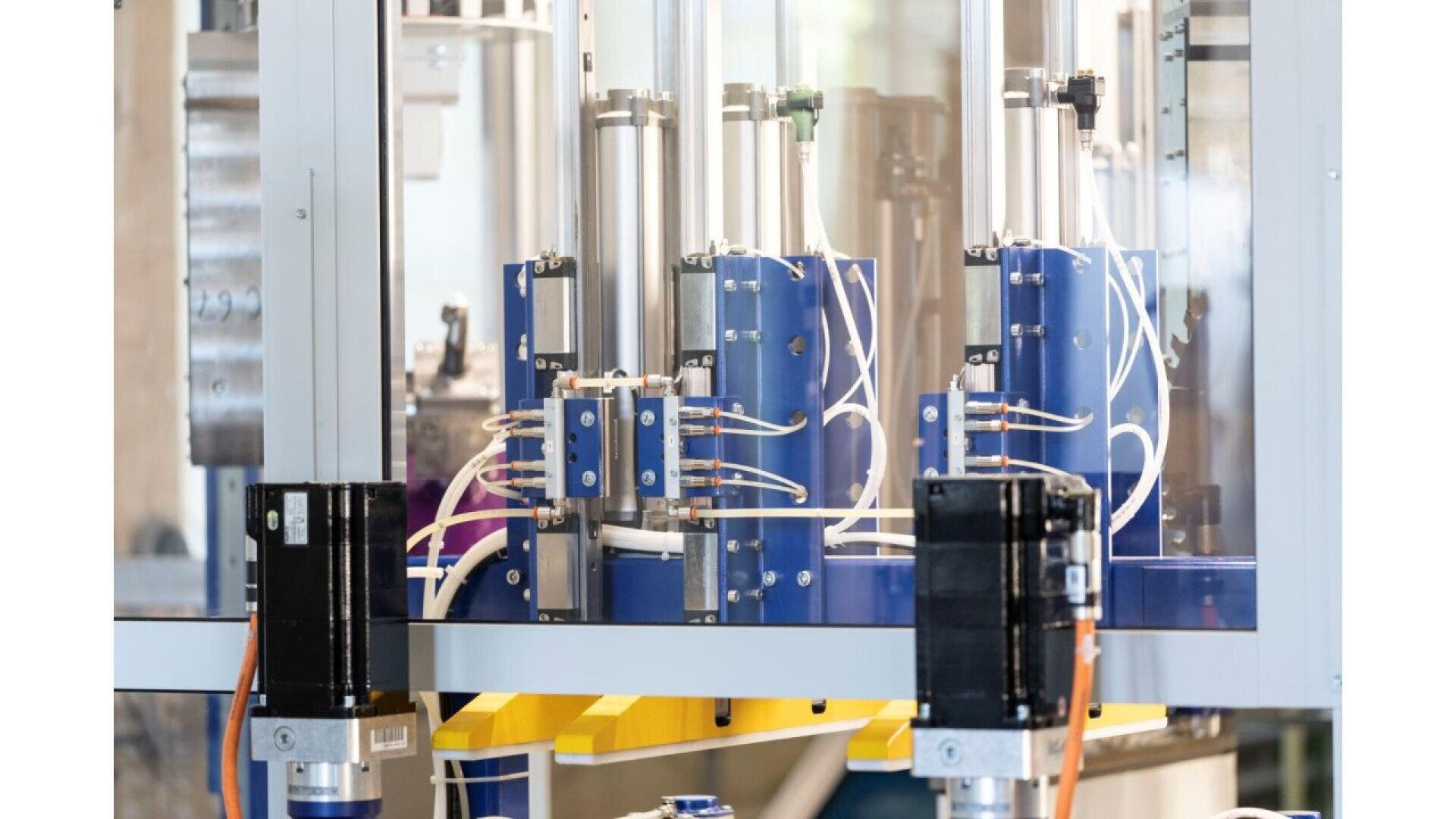 Gros plan d'un équipement d'automatisation industrielle comprenant des cylindres, des tubes et des fils pneumatiques bleus et argentés à l'intérieur d'une enceinte en verre, avec des composants noirs et orange au premier plan.