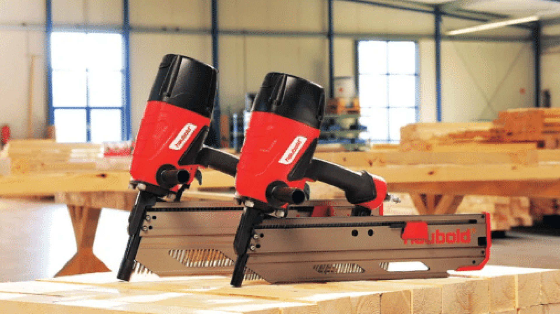Zwei rot-schwarze Druckluftnagler stehen auf einer Holzfläche in einer Werkstatt, in der im Hintergrund Holzstapel und Werkzeuge zu sehen sind.