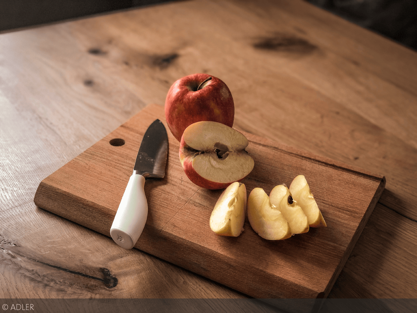 Ein roter Apfel, ein Messer mit weißem Griff und mehrere Apfelscheiben liegen auf einem hölzernen Schneidebrett auf einem Holztisch.