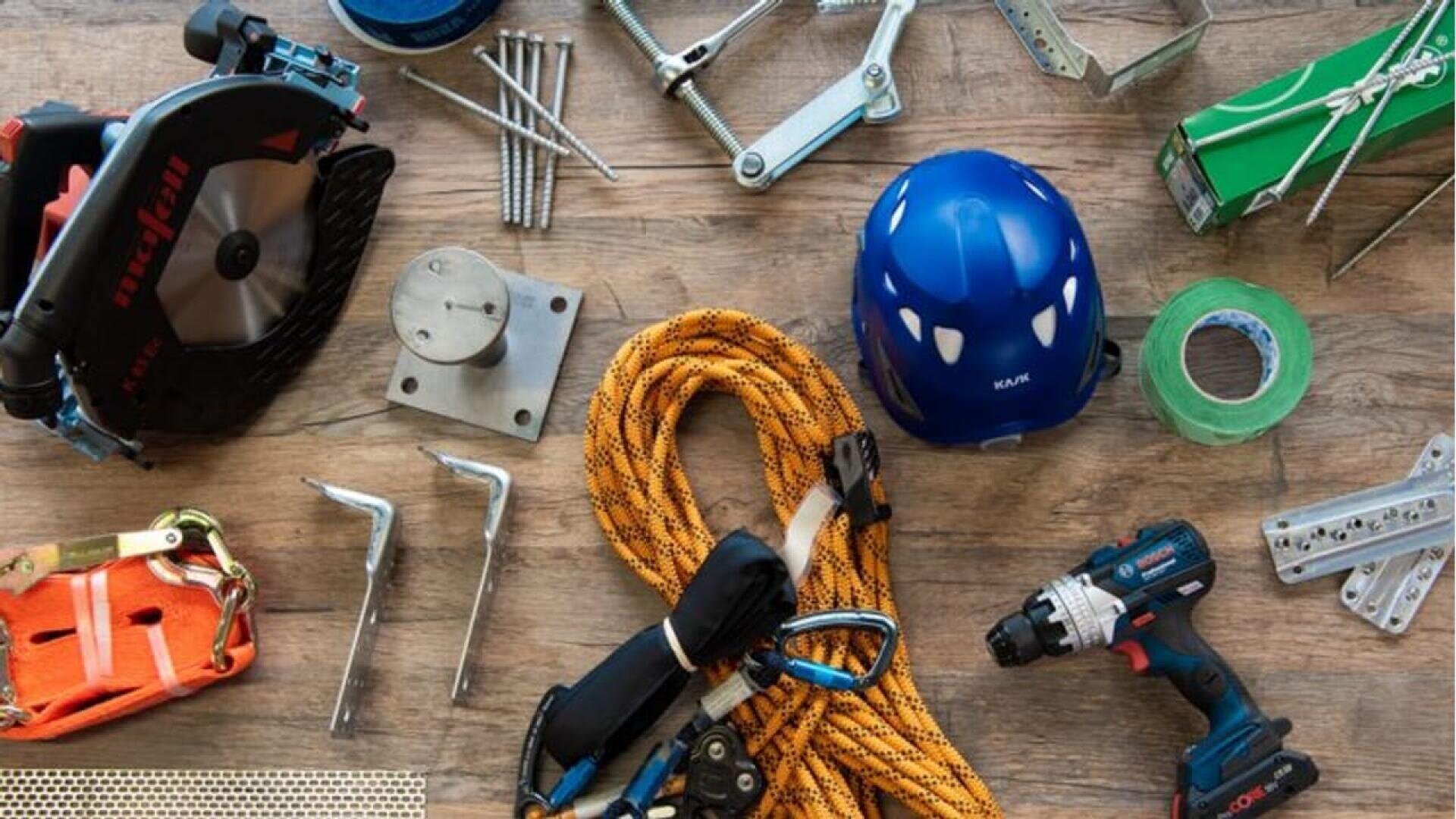 Divers outils de construction et équipements de sécurité disposés sur une surface en bois, notamment une scie circulaire, un casque, une perceuse, une corde, du ruban adhésif, des courroies, des supports métalliques, des vis et des boulons.