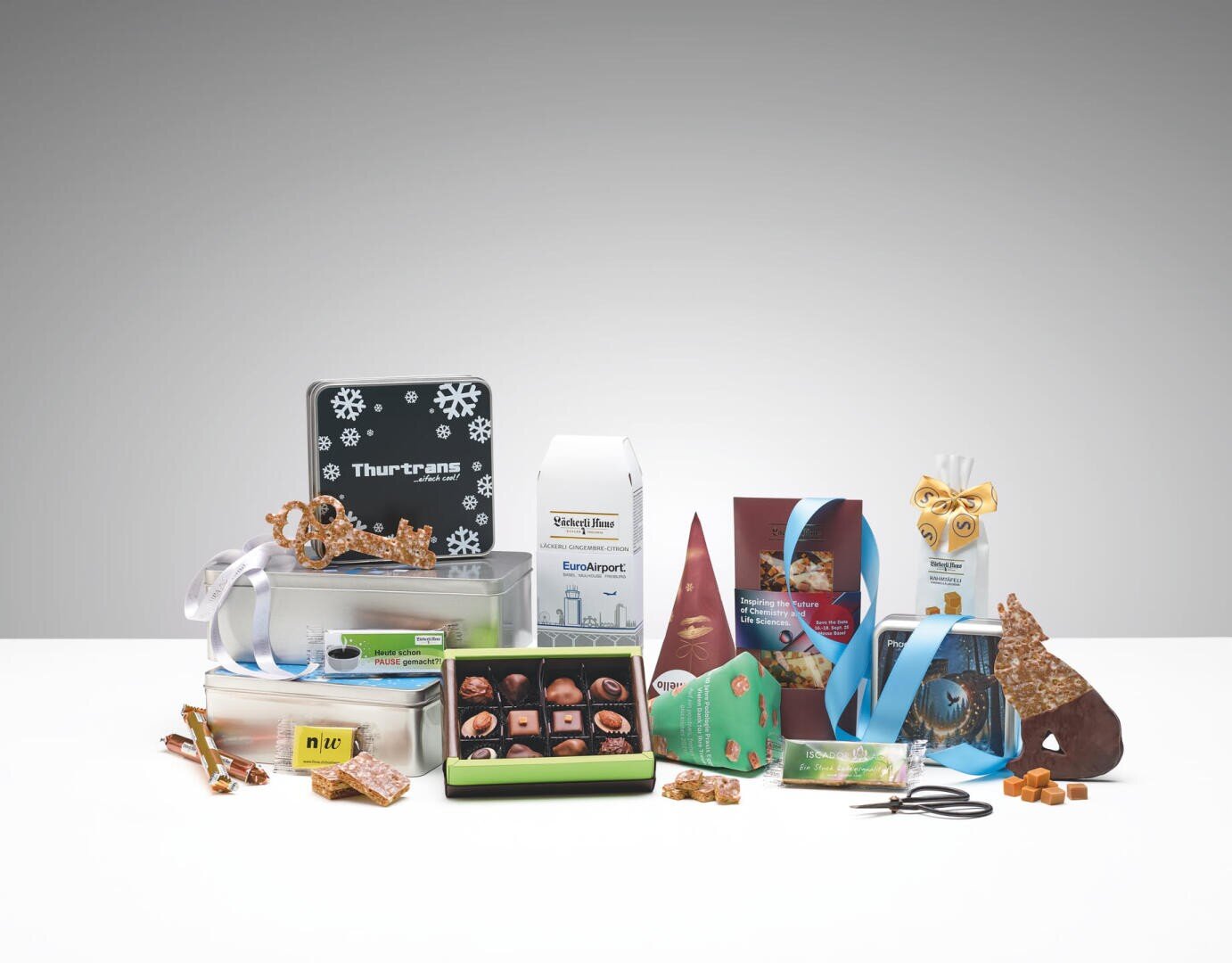 Eine Vielzahl von verschiedenen Süßigkeiten und Snacks, Schokolade, Gebäck und Keksen sind auf einer weißen Fläche mit festlichen Verpackungen und Etiketten angeordnet. Einige Schachteln sind offen, um die Pralinen zu präsentieren; Dekorationen und Bänder sind enthalten.