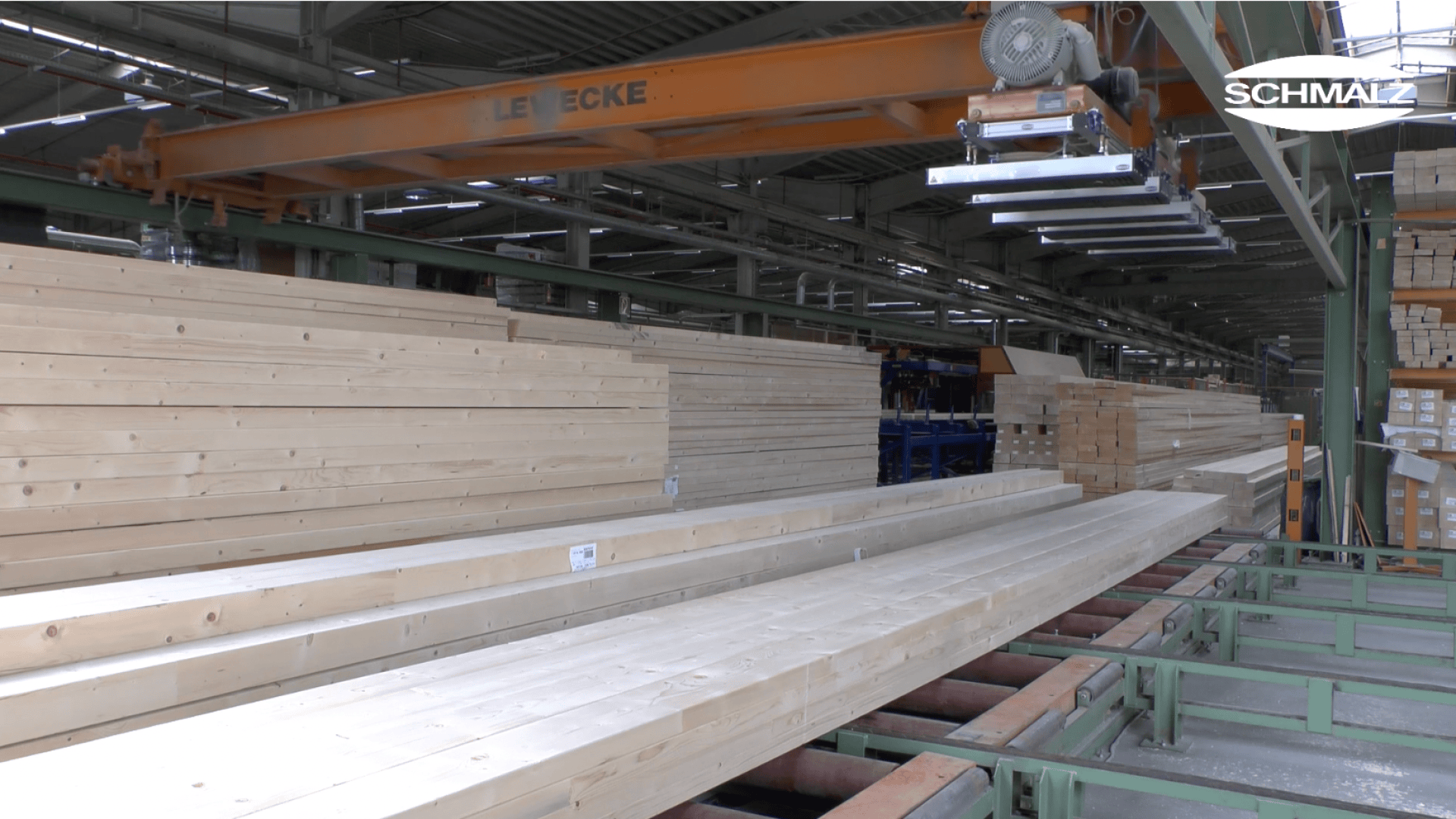 Ein Roboterarm mit einem Vakuum-Hebesystem bewegt lange Holzbalken in einem großen Industrielager. In der gut beleuchteten Anlage sind Stapel von Schnittholz aufgereiht. Das SCHMALZ-Logo erscheint in der oberen rechten Ecke.