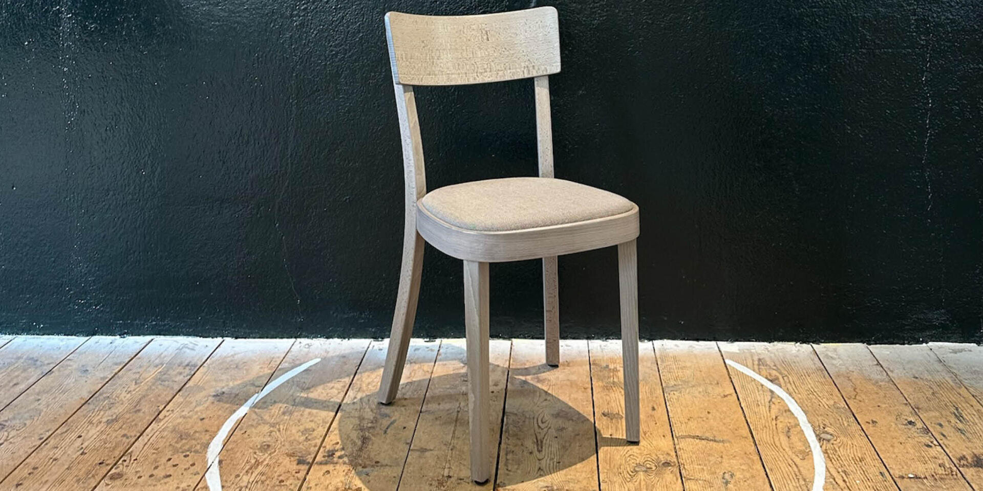 Une chaise en bois clair au dossier incurvé et à l'assise rembourrée est placée au centre d'un cercle blanc sur un sol en bois devant un mur noir.