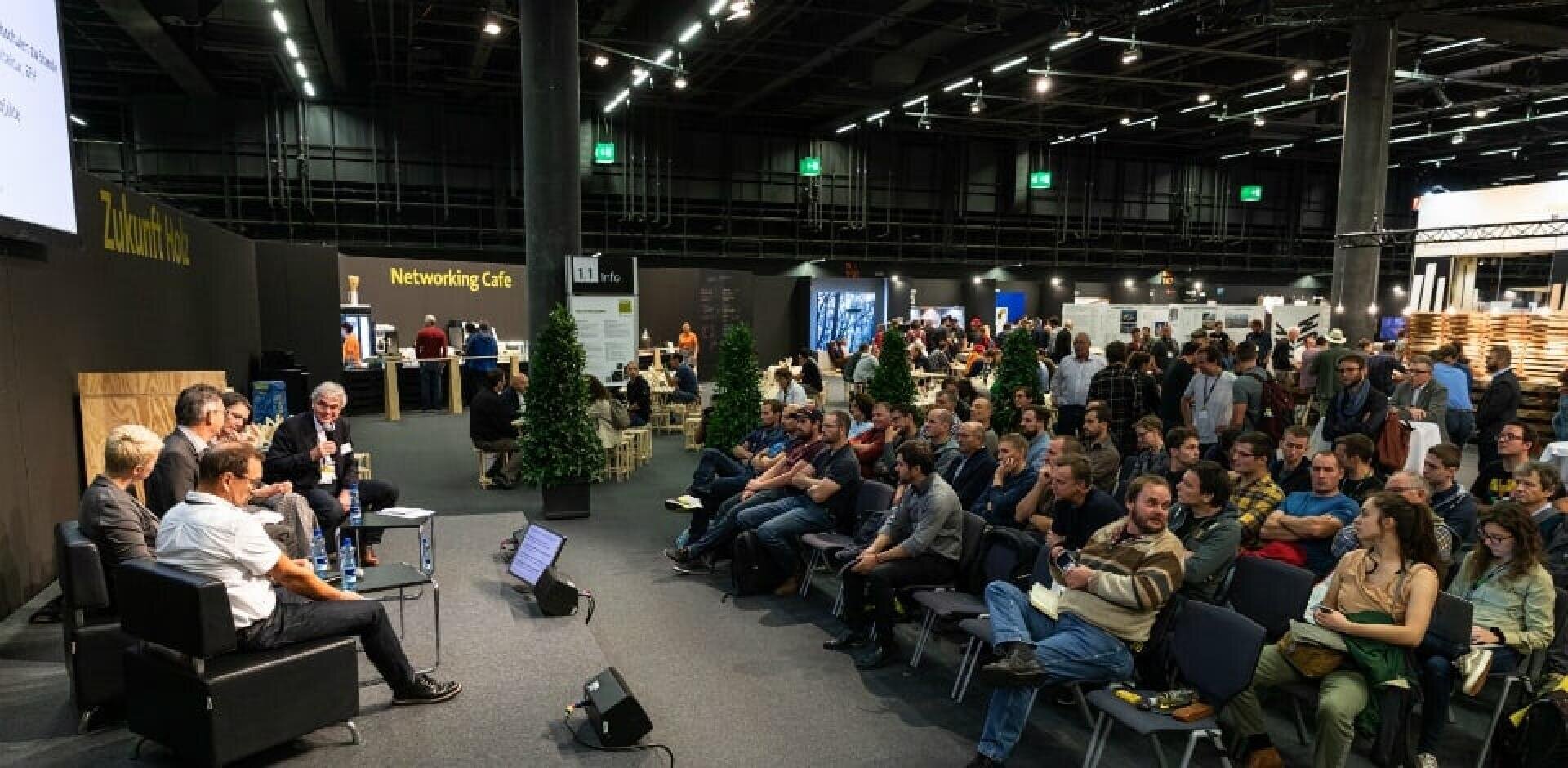Auf einer Konferenz sitzt ein großes Publikum vor einer Reihe von Rednern auf einer Bühne. Der Raum ist geräumig, mit hohen Decken, verstreutem Grün und Schildern für ein Networking Café und verschiedene Stände im Hintergrund.