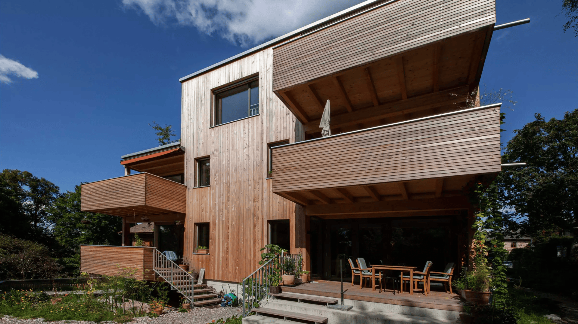 Ein modernes dreistöckiges Haus mit einem Außenbereich aus Holz, großen Balkonen und einem Esszimmer im Freien. Das Gebäude ist von viel Grün umgeben und der Himmel ist von einem klaren Blau.