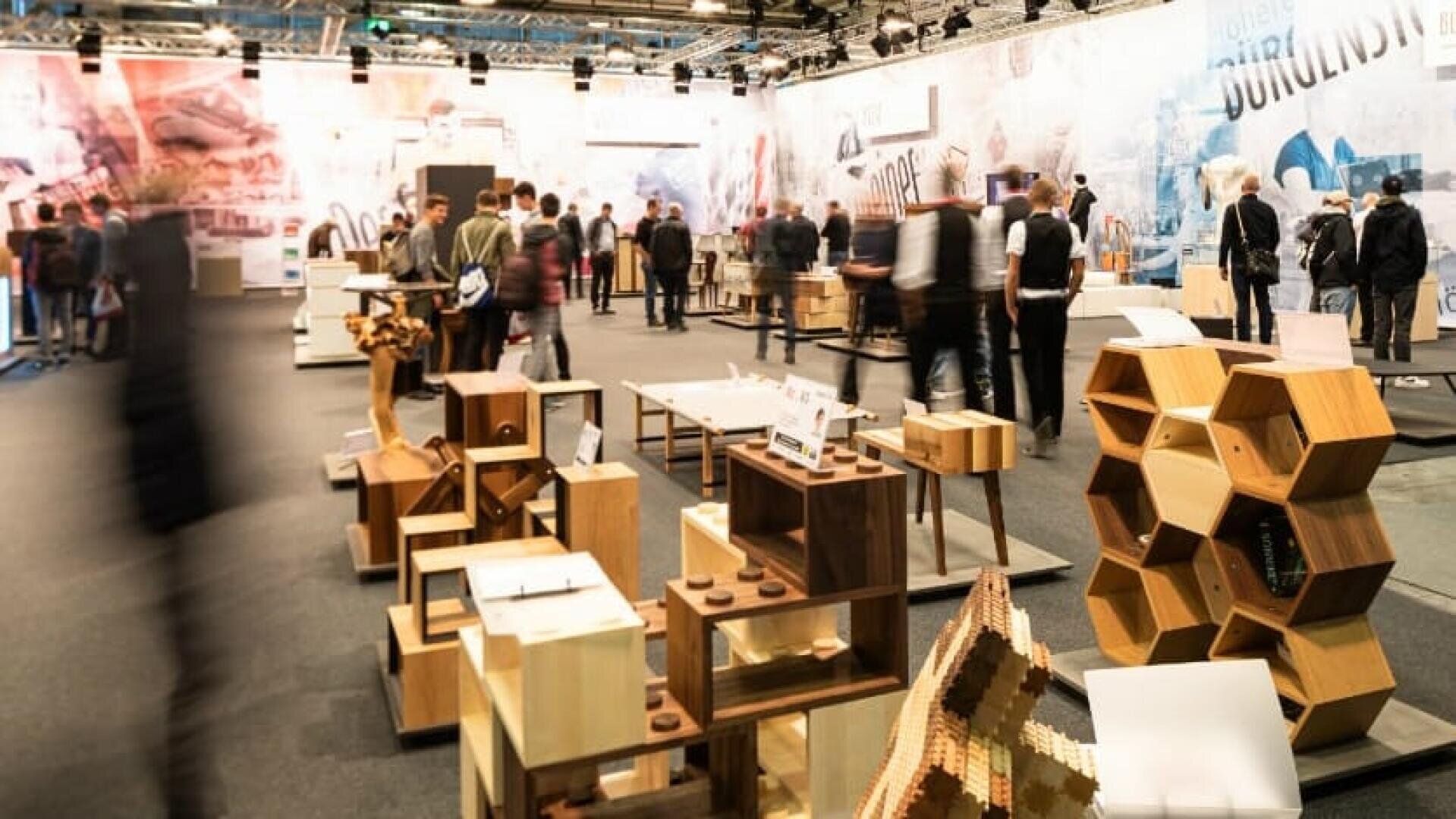 Die Besucher gehen durch eine Innenausstellung mit modernen Holzmöbeln und geometrischen Regalen. Der Raum ist hell erleuchtet, mit Displays und Informationsschildern im gesamten Bereich.