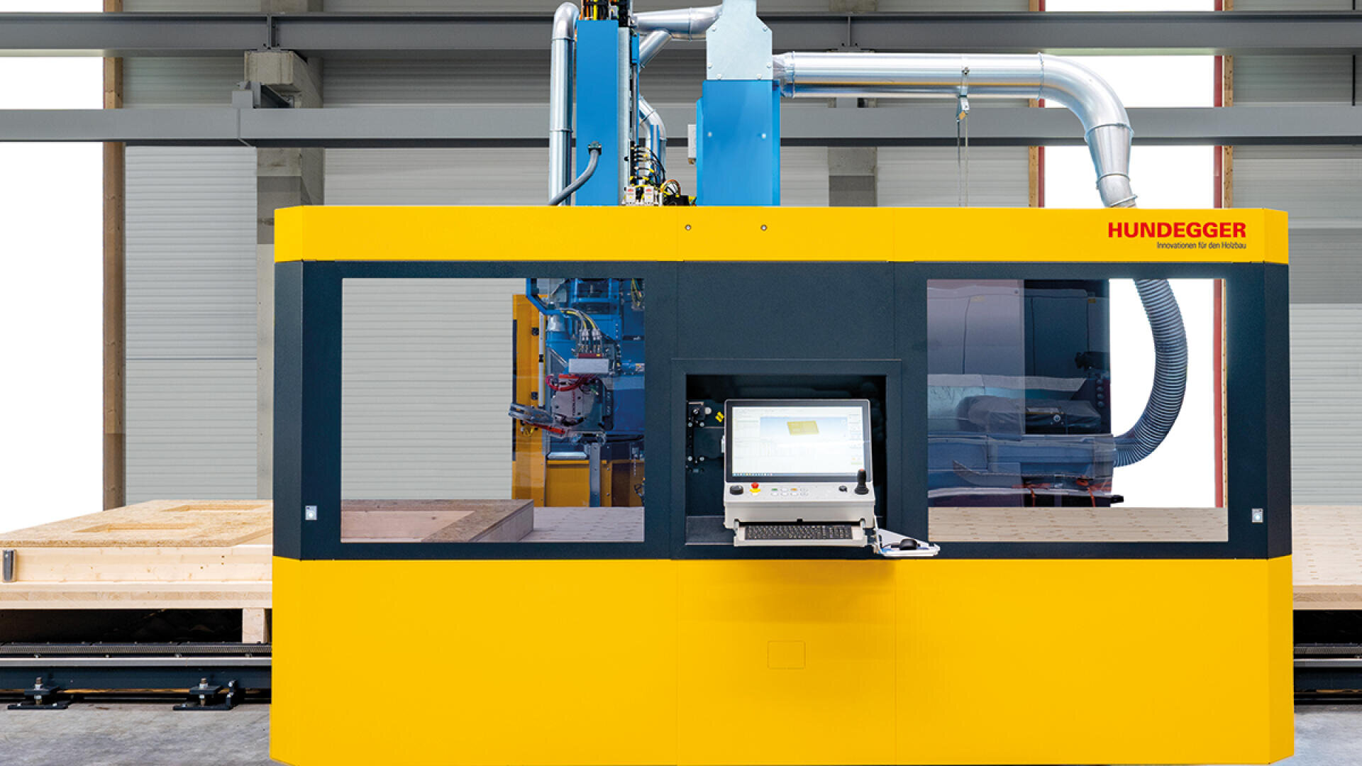 Eine gelbe und blaue Hundegger-Industrieholzbearbeitungsmaschine mit einem Touchscreen-Bedienfeld ist in einer Fabrikhalle aufgestellt, umgeben von großen Holzbalken und modernen Geräten.