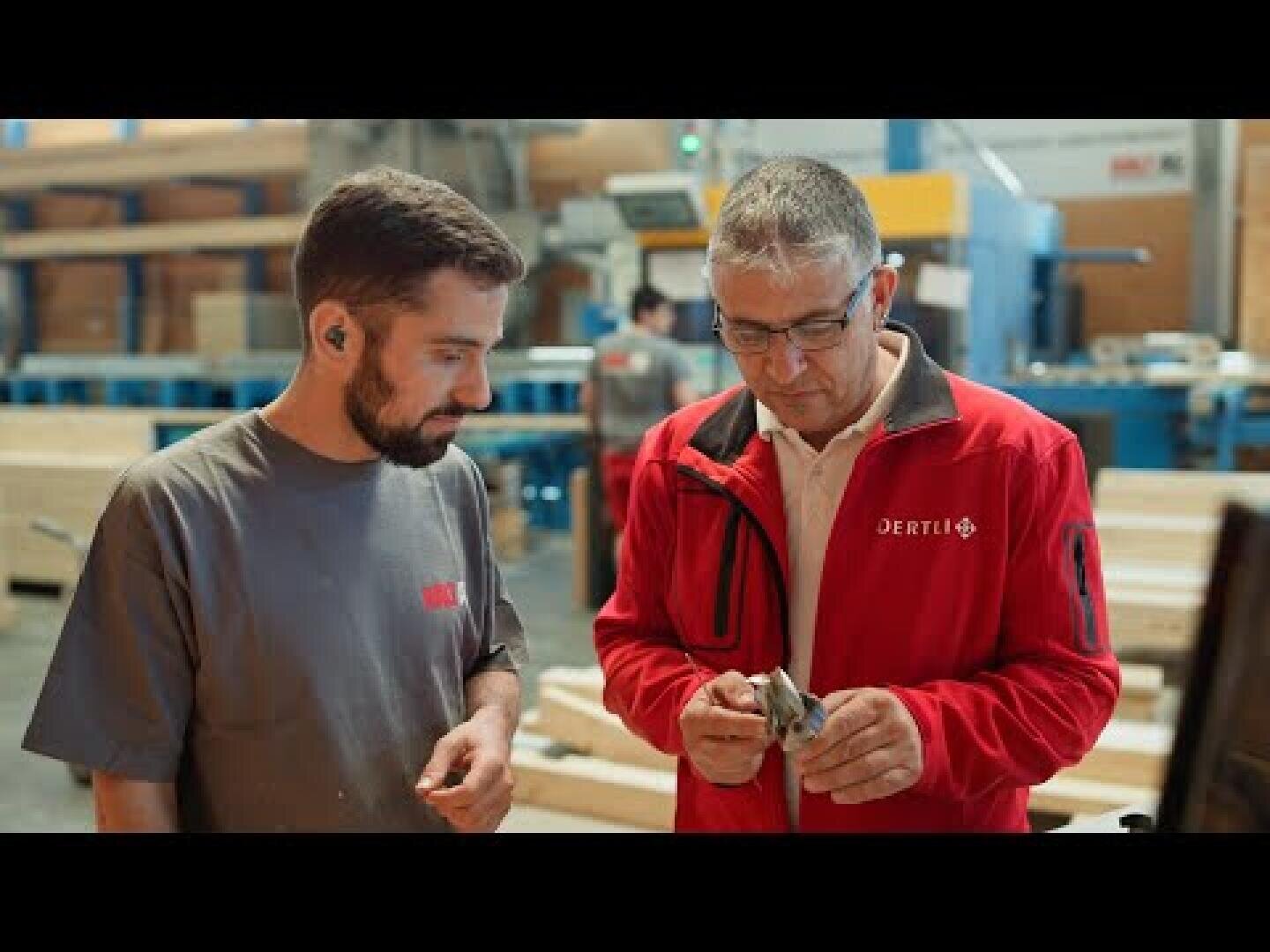 Zwei Männer stehen in einer Werkstatt. Der eine trägt ein graues Hemd und hört aufmerksam zu, während der andere in einer roten Jacke ein kleines Metallteil inspiziert. Im Hintergrund sind Maschinen und Holzmaterialien zu sehen.