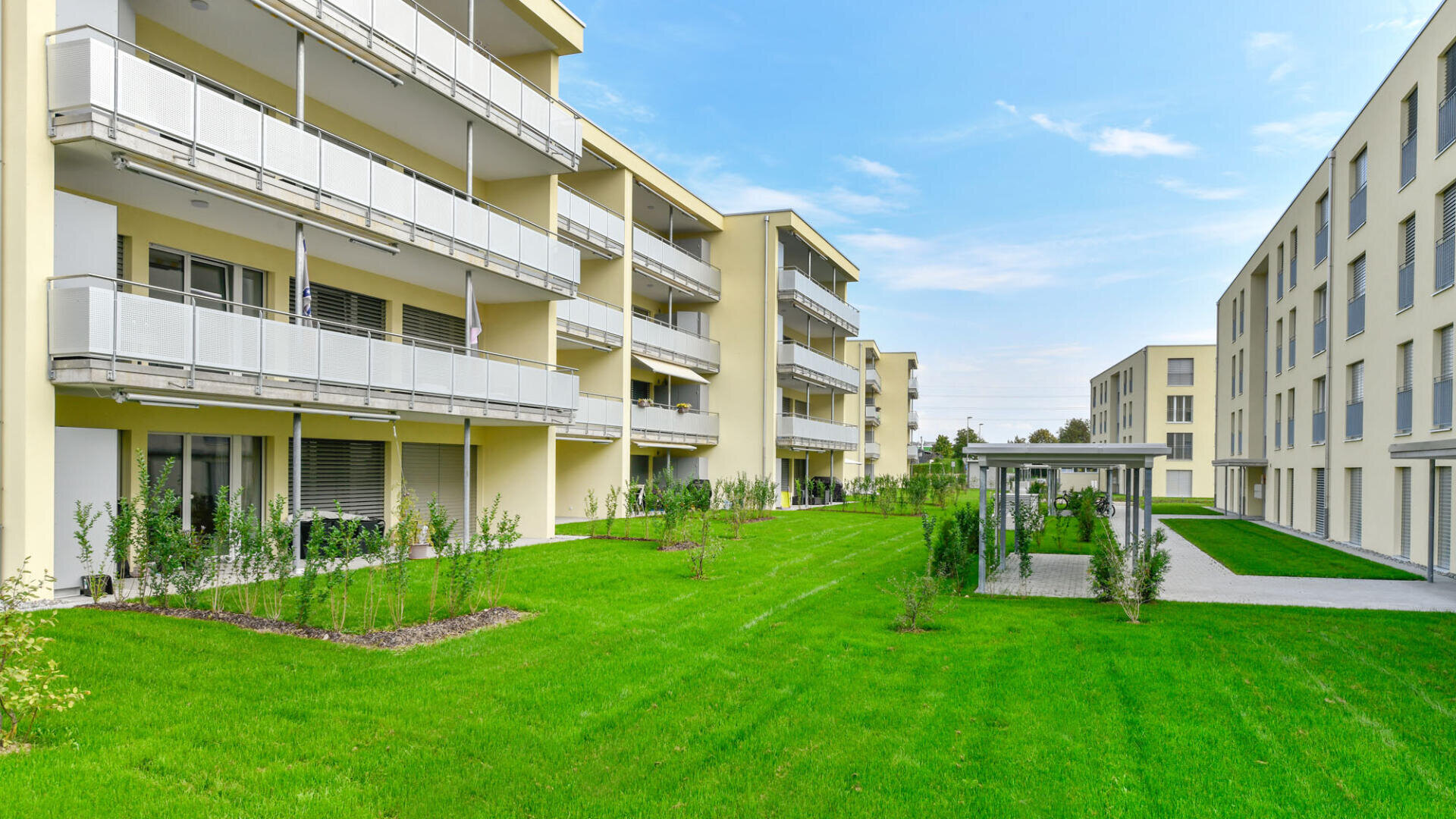 Moderne Mehrfamilienhäuser mit Balkonen stehen vor einem gepflegten grünen Rasen und jungen Sträuchern unter blauem Himmel, mit einem überdachten Gehweg zwischen den beiden Gebäuden.
