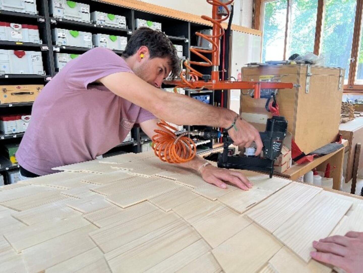 Dans un atelier, une personne portant des bouchons d'oreille travaille de fines plaques de bois avec un outil pneumatique. En arrière-plan, on voit des étagères avec des caisses bien rangées et une grande fenêtre.