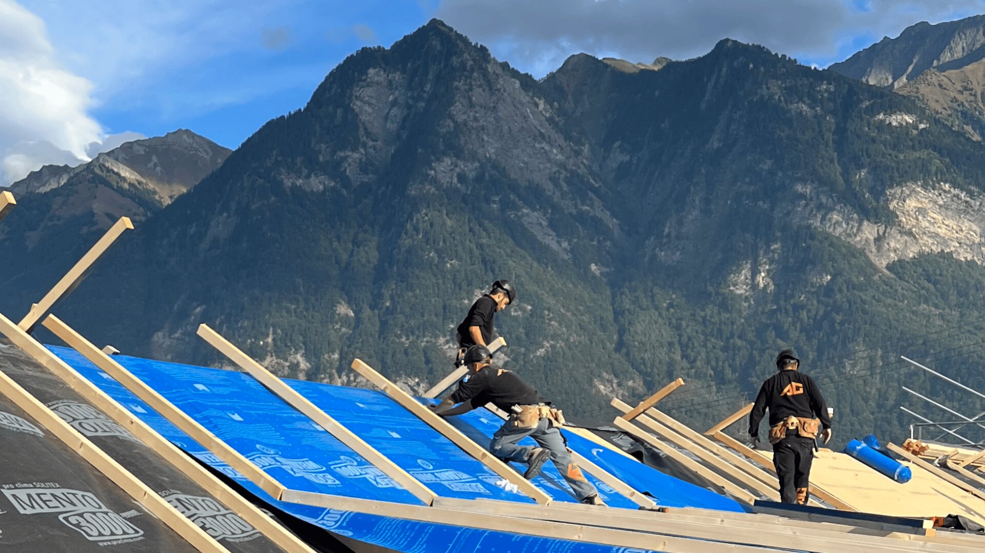 Drei Arbeiter installieren unter freiem Himmel eine blaue Isolierung auf einem Schrägdach, im Hintergrund sind hohe, bewaldete Berge zu sehen.