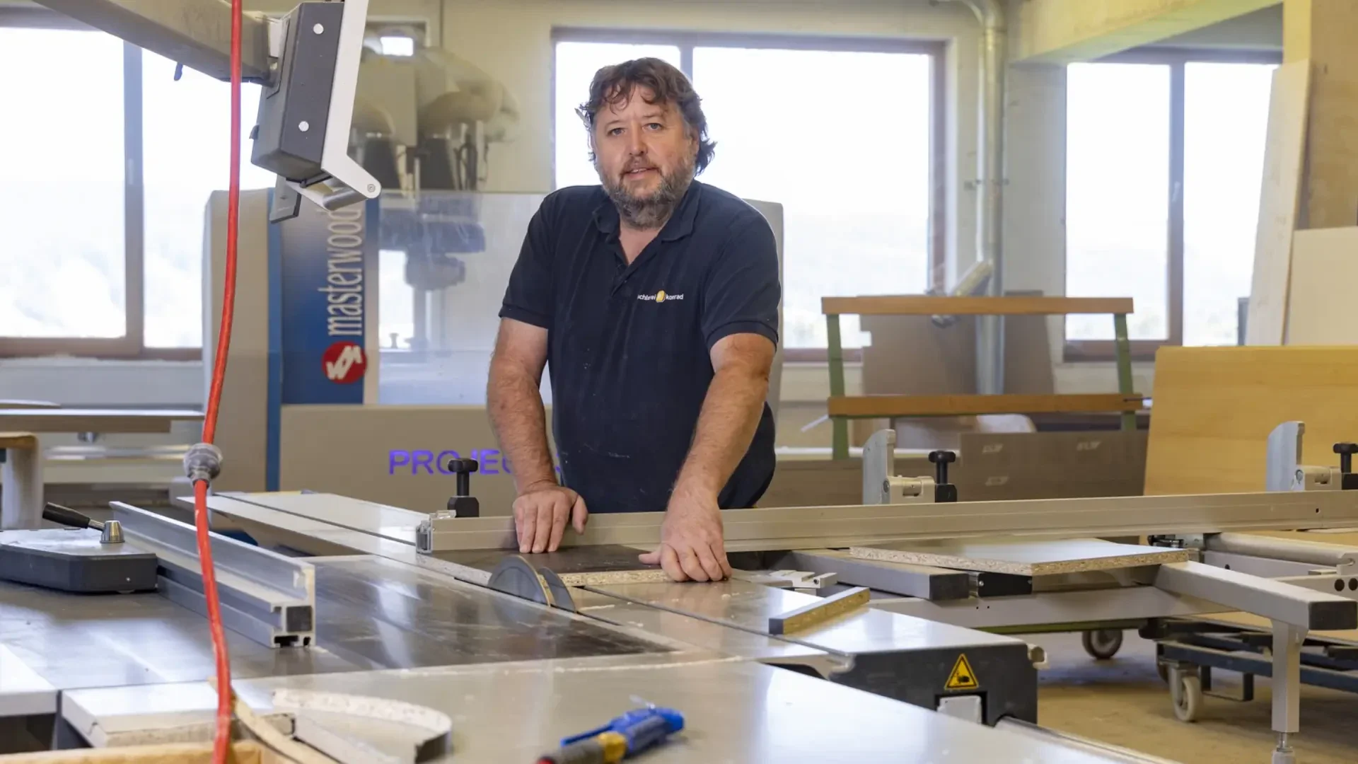Ein Mann steht in einer Holzwerkstatt und bedient eine Tischsäge, wobei er das Material mit den Händen berührt. Um ihn herum sind verschiedene Holzbearbeitungsgeräte und Werkzeuge in dem gut beleuchteten Arbeitsbereich zu sehen.