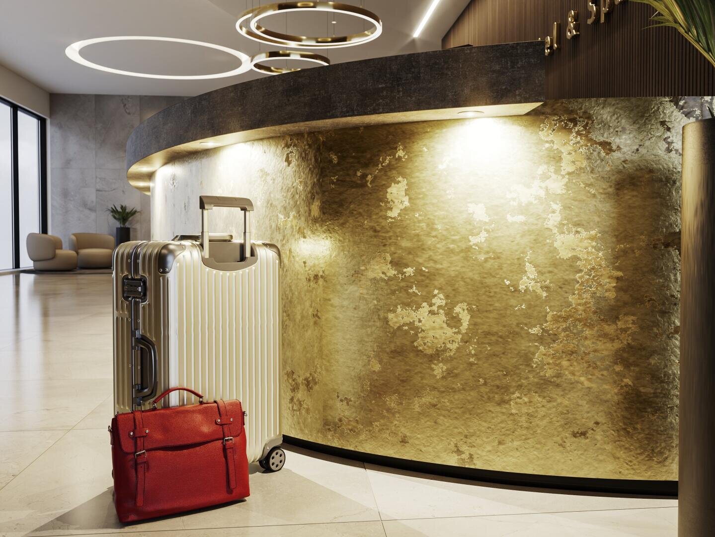 Une valise argentée et un attaché-case rouge sont posés devant la réception dorée d'un hôtel, dans un hall moderne et bien éclairé.