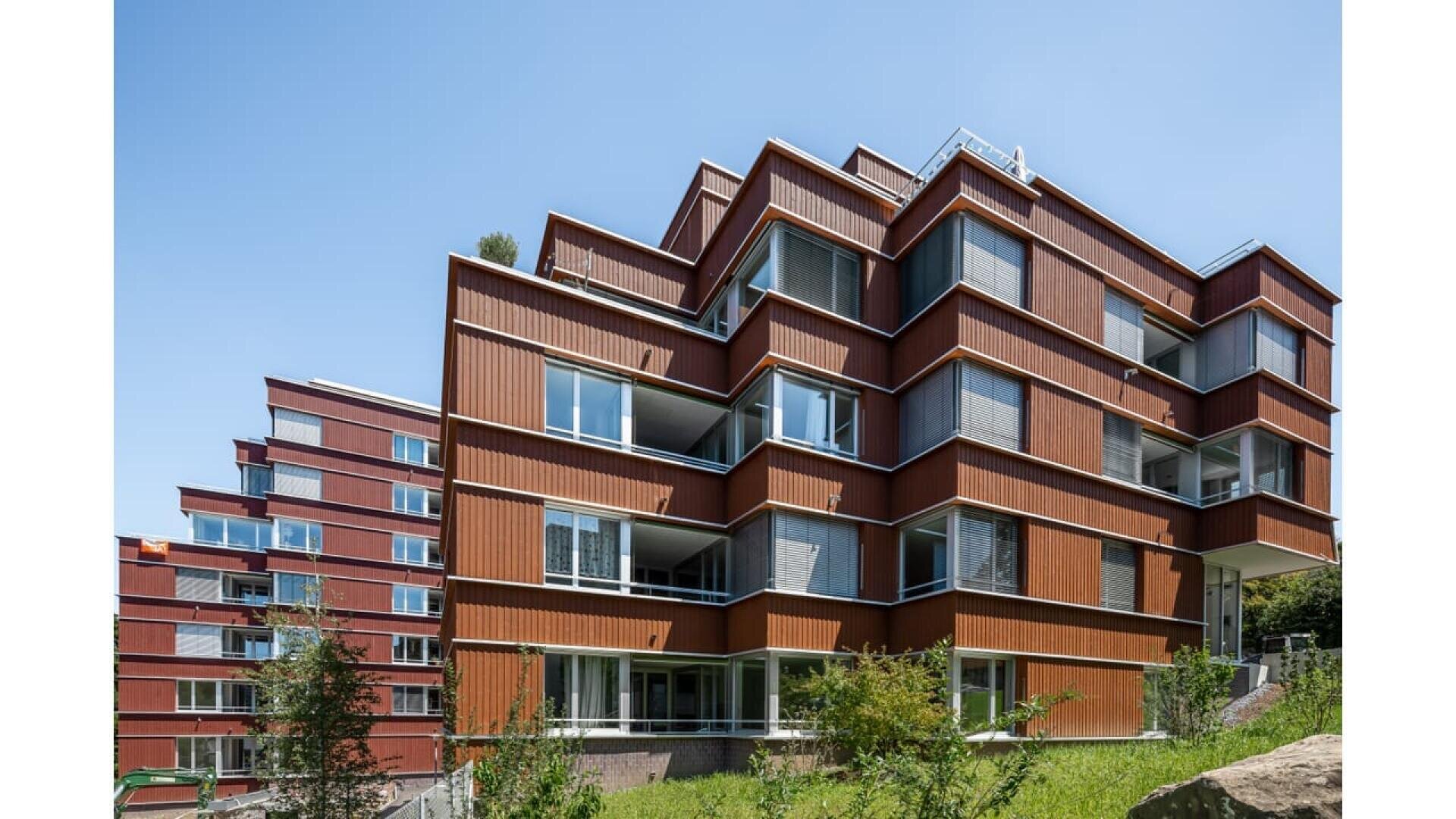 Immeubles d'habitation modernes avec des façades rouge-brun en quinconce, de grandes fenêtres et des garde-corps blancs, sur fond de ciel bleu clair et de verdure au premier plan.