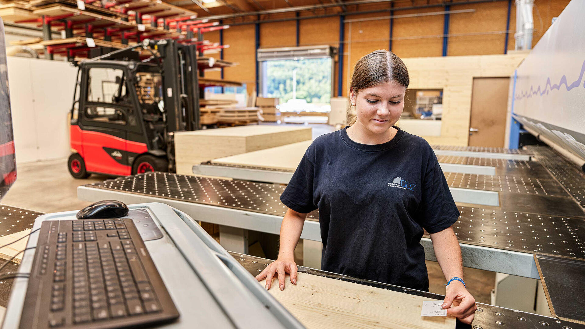 Eine Frau in einem dunklen T-Shirt bedient Maschinen in einer Holzwerkstatt und hält eine Karte neben einem Scanner. Eine Computertastatur und eine Maus befinden sich an einem nahe gelegenen Arbeitsplatz, und im Hintergrund ist ein Gabelstapler zu sehen.