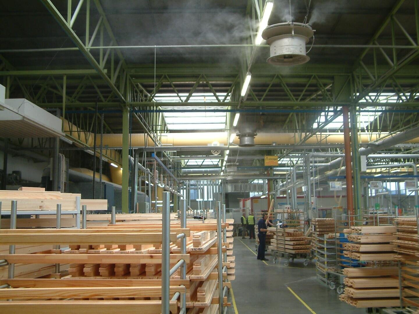 Ein geräumiges, gut beleuchtetes Fabrikinnere mit Stapeln von Holzbrettern auf Metallregalen, einer hohen Decke mit Balken und Arbeitern im Hintergrund. In der Nähe des Daches sind ein Ventilator und Nebel zu sehen.