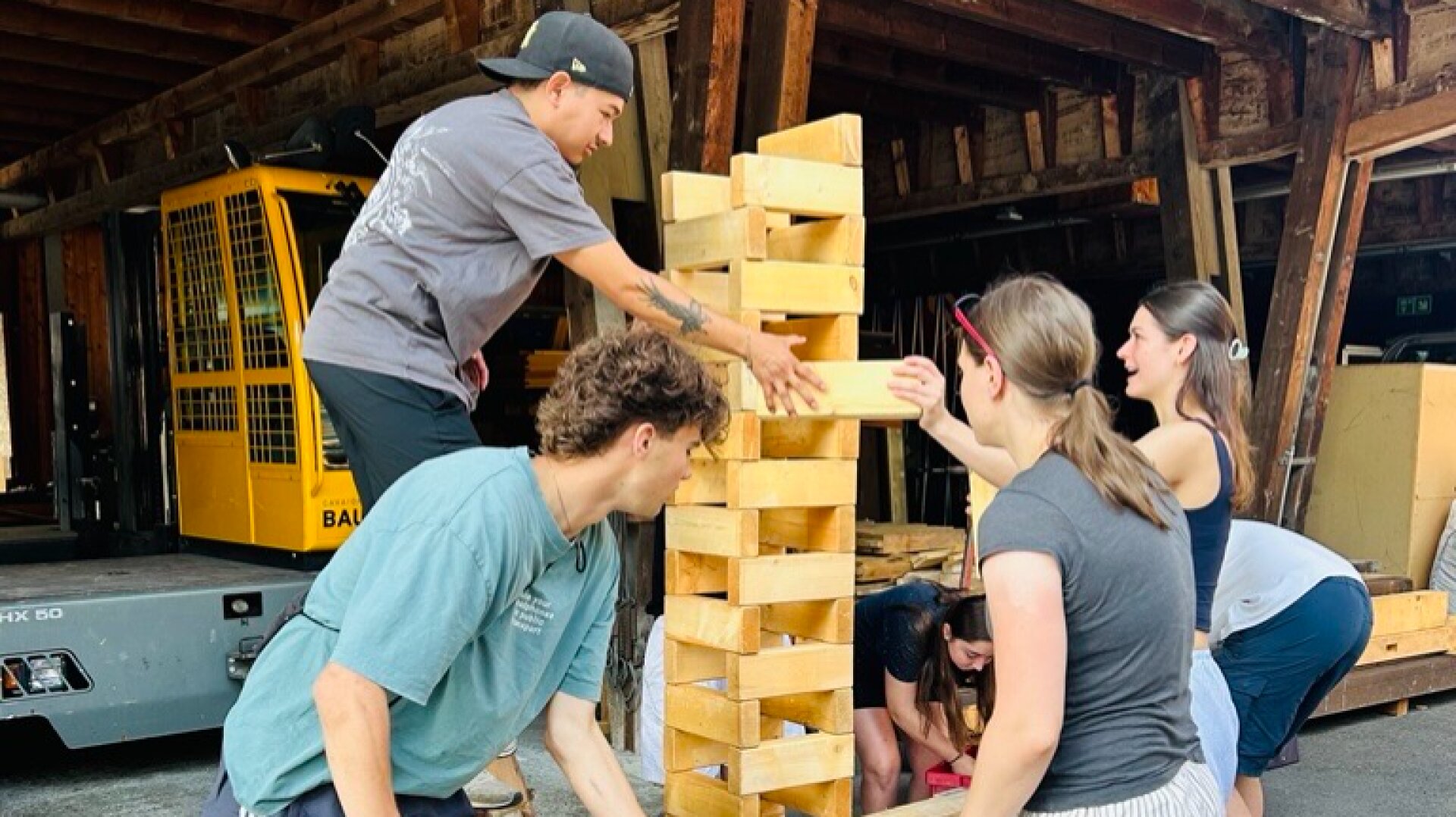 Fünf Personen spielen in einer Garage oder einem Lagerhaus ein überdimensionales Jenga-Spiel aus Holz. Sie konzentrieren sich auf das sorgfältige Entfernen und Stapeln von Blöcken, wobei im Hintergrund ein Gabelstapler und Holzbalken zu sehen sind.