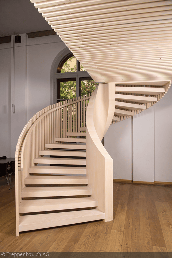 Eine moderne, helle Holzspindeltreppe mit vertikalen Latten und sanften Kurven steht in einem Raum mit großen Fenstern, weißen Wänden und einem Holzboden. Das einfallende Sonnenlicht bringt die Handwerkskunst der Treppe zur Geltung.