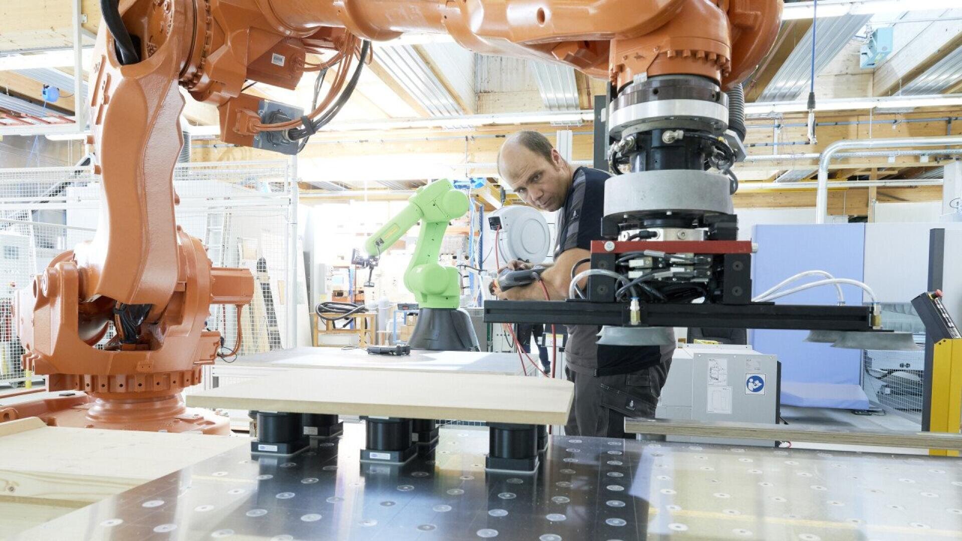 Un homme manipule un grand bras robotisé industriel dans un atelier moderne, avec un autre bras robotisé vert et diverses machines visibles à l'arrière-plan. Il se concentre sur le réglage de l'équipement.