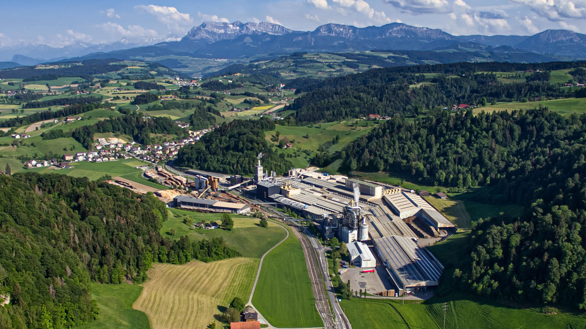 Luftaufnahme eines Industriekomplexes, umgeben von grünen Feldern, Wäldern und kleinen Dörfern, mit Bergen im Hintergrund unter einem teilweise bewölkten Himmel.