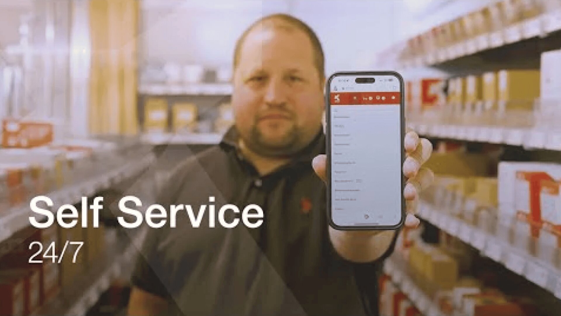 Ein Mann steht in einem Lagerraum und hält ein Smartphone in die Kamera, auf dem eine Selbstbedienungs-App angezeigt wird. Der Text auf dem Bild lautet: "Self Service 24/7". Regale mit Kisten füllen den Hintergrund.