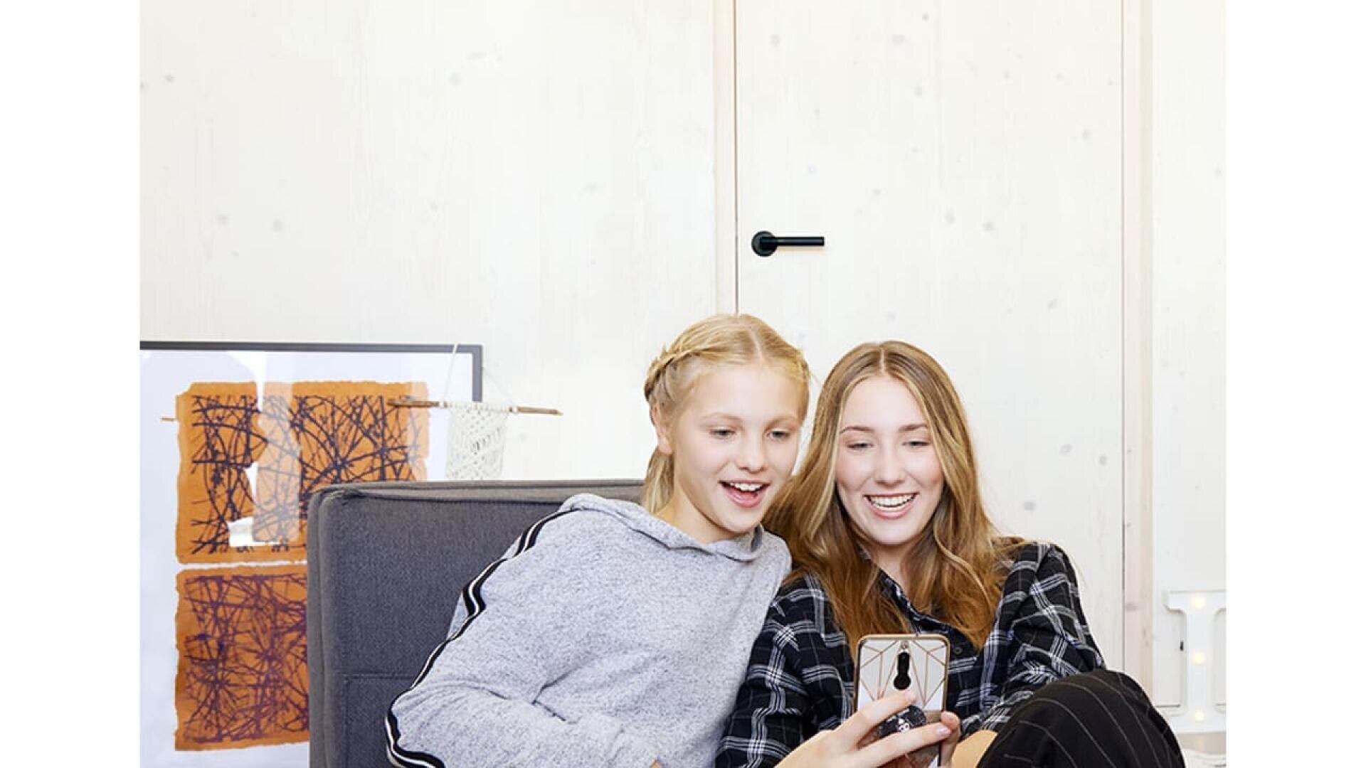 Deux filles sont assises ensemble sur un canapé, souriant et regardant un smartphone que l'une d'entre elles tient à la main. Elles semblent apprécier quelque chose à l'écran dans une pièce lumineuse et confortable au décor minimal.