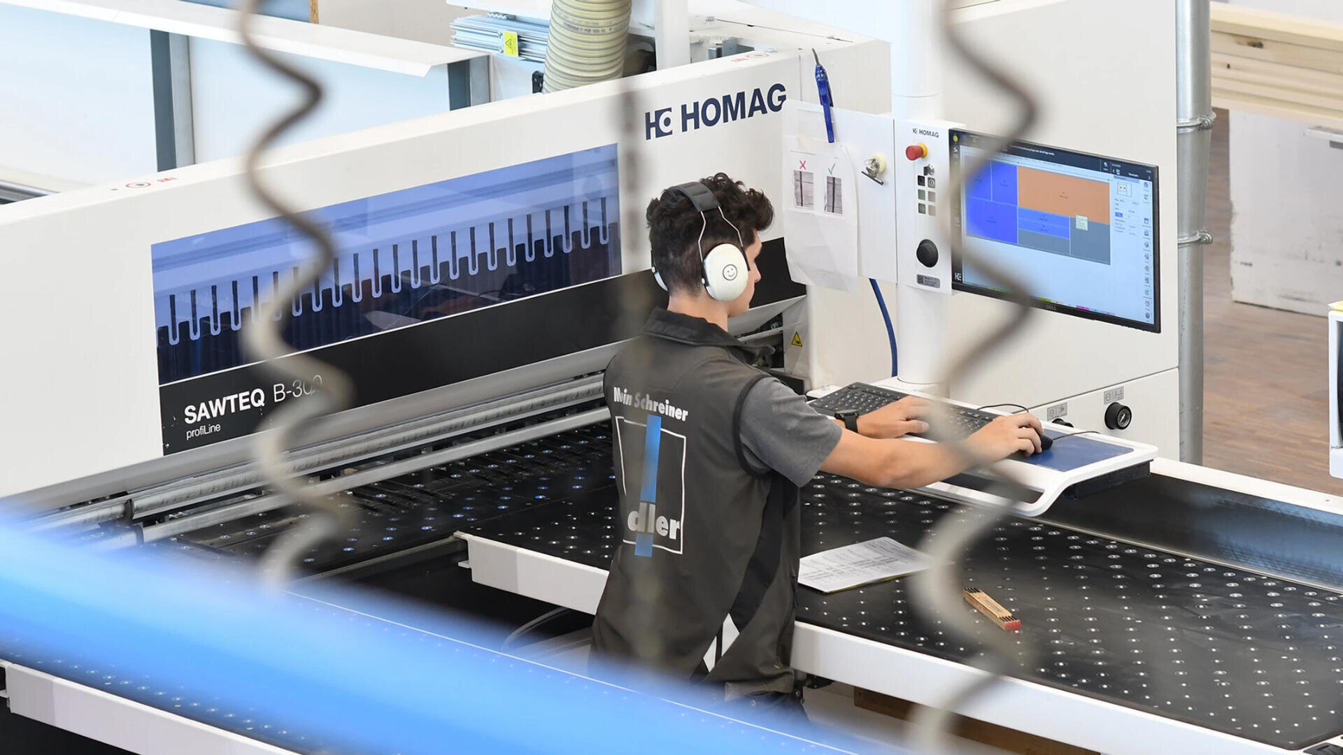 Eine Person mit Kopfhörern bedient einen Computer an einer modernen Industriemaschine in einer Fabrik. Auf dem Bildschirm wird eine farbcodierte Schnittstelle angezeigt, und der Arbeiter trägt eine Weste mit Firmenlogo.