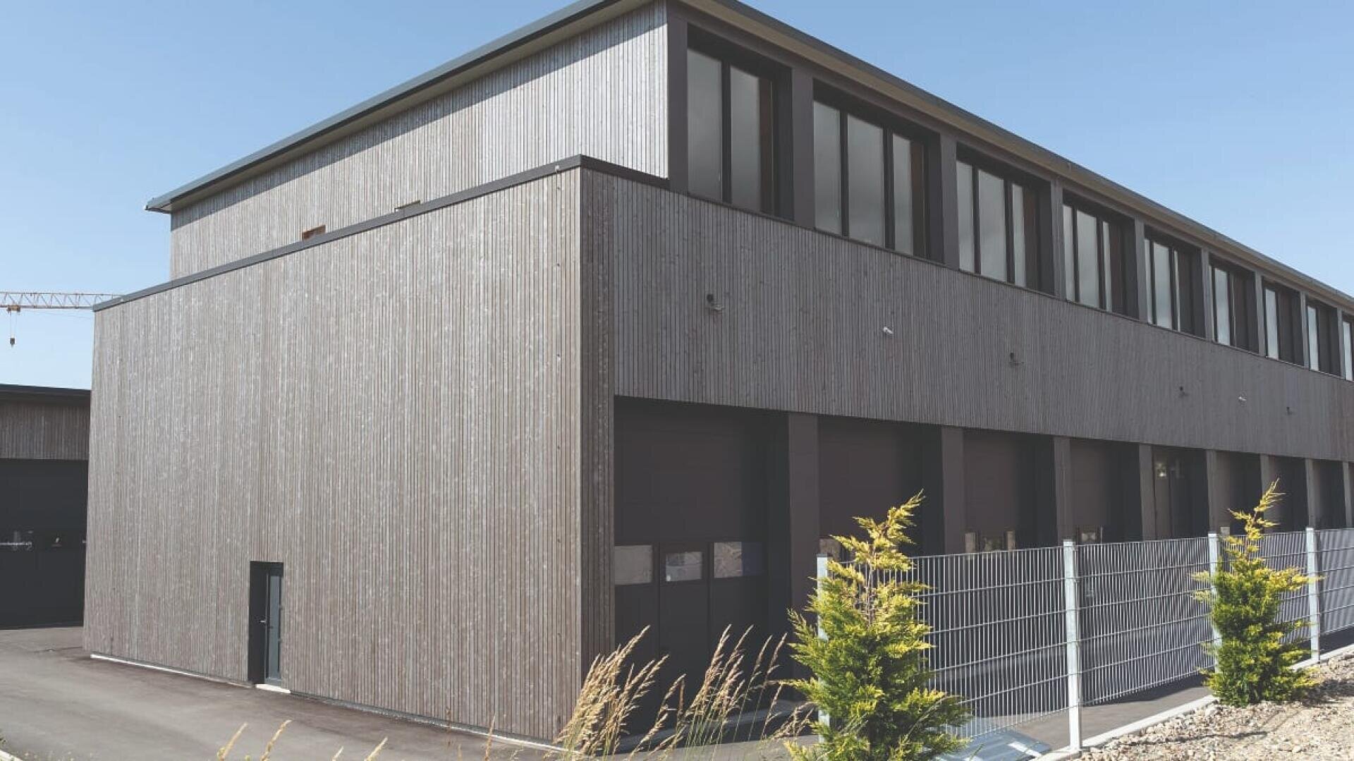Ein modernes Industriegebäude mit einer grauen Holzfassade, großen Fenstern im Obergeschoss und einem Metallzaun davor. Ein paar kleine immergrüne Sträucher sind in der Nähe des Zauns gepflanzt. Der Himmel ist klar und blau.