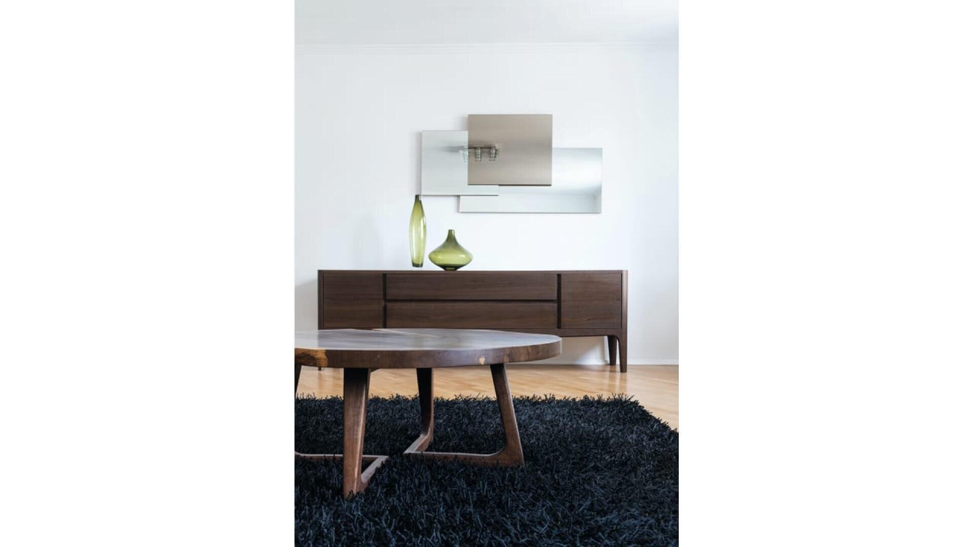 Salon moderne avec une table basse en bois foncé sur un tapis noir en shaggy, un buffet en bois avec un vase vert, et des miroirs abstraits sur un mur blanc.