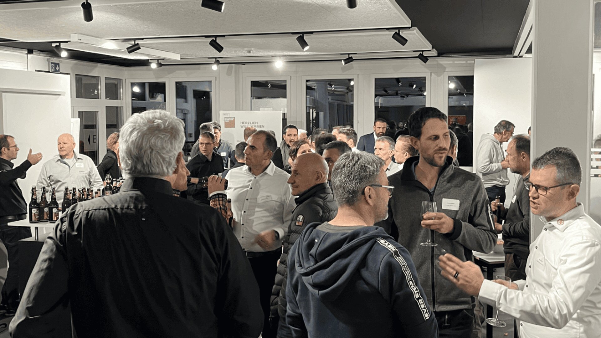 Un grand groupe de personnes en tenue d'affaires discutent lors d'un événement de réseautage en intérieur. Certains tiennent des boissons et des bouteilles sont posées sur une table au premier plan. La salle est bien éclairée et le décor est moderne.