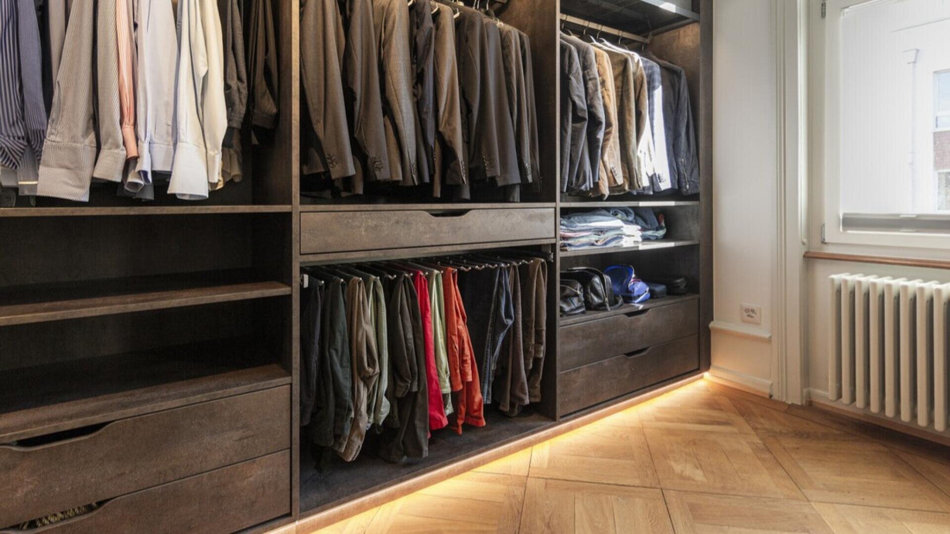 Un dressing bien organisé avec des étagères, des tiroirs et un espace de suspension pour les costumes, les chemises, les pantalons et les vêtements pliés. La lumière naturelle pénètre par une fenêtre voisine sur un sol en parquet.
