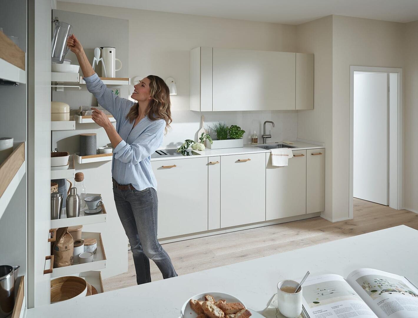 Eine Frau in einer modernen Küche greift mit einer Tasse in der Hand in ein hohes Regal. Die Küche besteht aus weißen Schränken, Holzregalen und einer Arbeitsplatte mit einem aufgeschlagenen Buch, einem Teller mit Essen und einer Tasse.