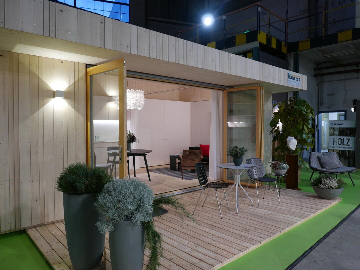 Ein modernes kleines Holzhaus mit großen Glastüren öffnet sich zu einer kleinen Terrasse mit Topfpflanzen und Sitzgelegenheiten im Freien. Das Innere ist sichtbar, mit minimalistischen Möbeln und einer Leuchte.