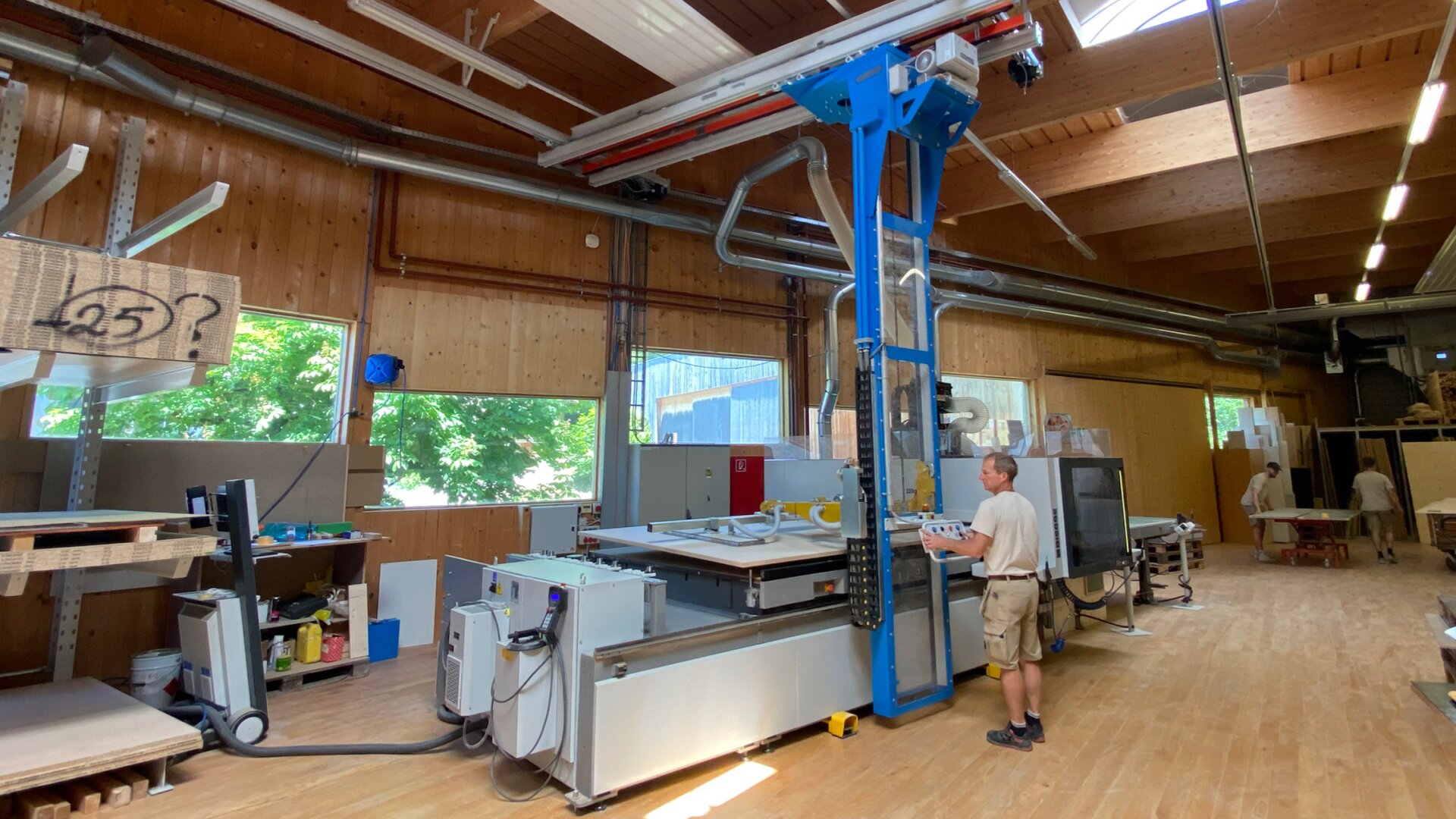 Eine Person bedient eine große, blaue automatische Maschine in einer geräumigen, gut beleuchteten Holzwerkstatt mit Holzwänden, hohen Decken und großen Fenstern. In der Werkstatt sind verschiedene Materialien und Geräte ordentlich angeordnet.