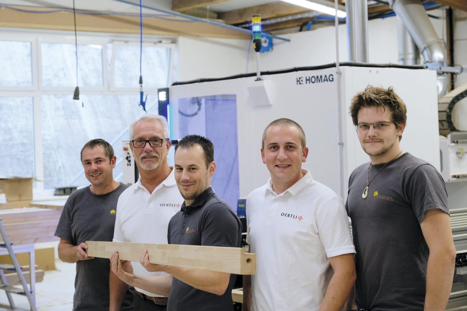 Fünf Männer stehen zusammen in einer Werkstatt und lächeln in die Kamera. Drei tragen passende weiße Polos, zwei tragen graue Polos, und einer hält ein Holzbrett. Im Hintergrund sind Werkstattausrüstung und Maschinen zu sehen.