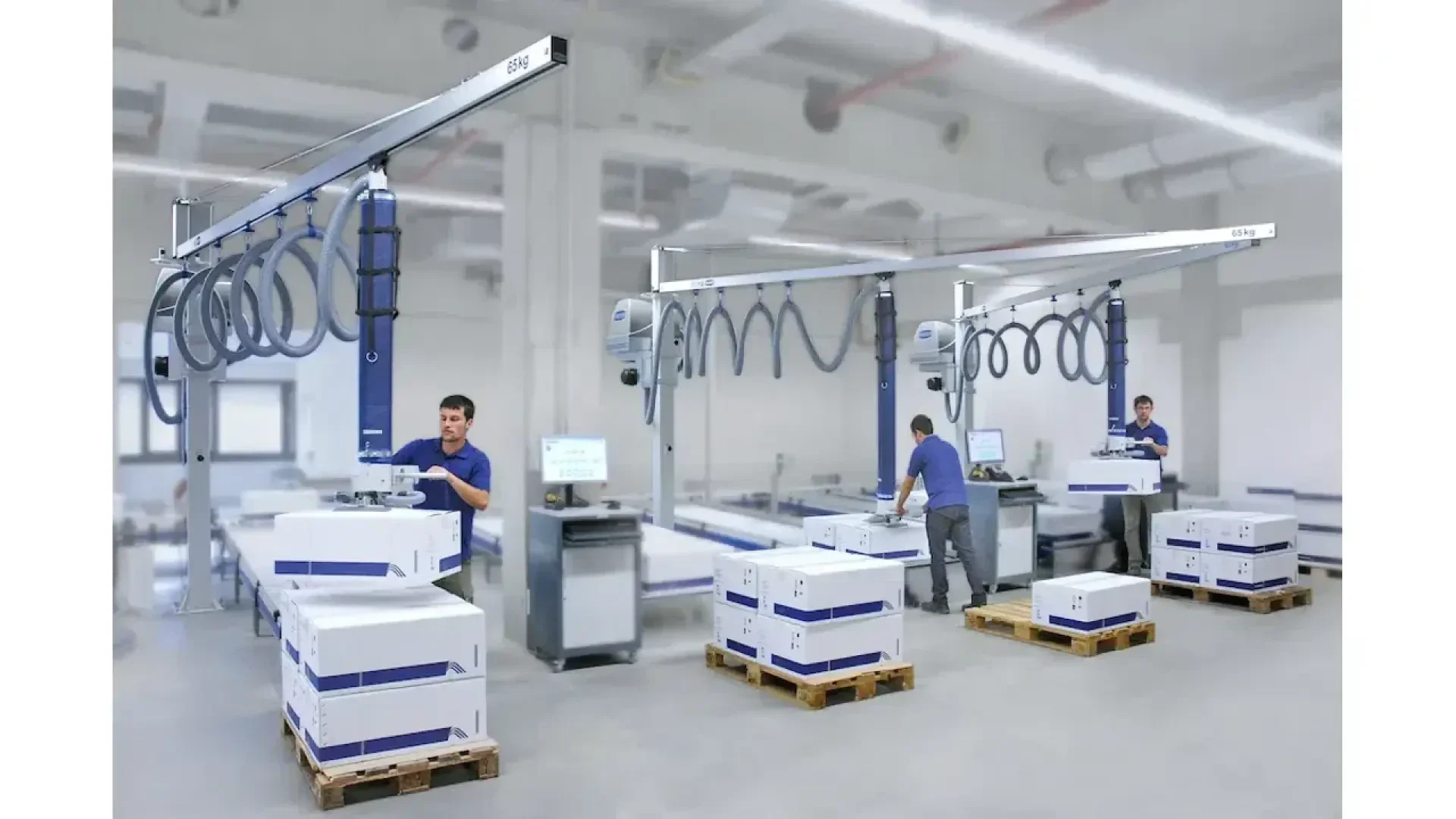 Dans un entrepôt moderne et lumineux, des travailleurs utilisent un équipement de levage aérien pour déplacer de grandes boîtes blanches sur des palettes en bois. Des postes informatiques et diverses machines sont visibles dans cet espace propre et organisé.