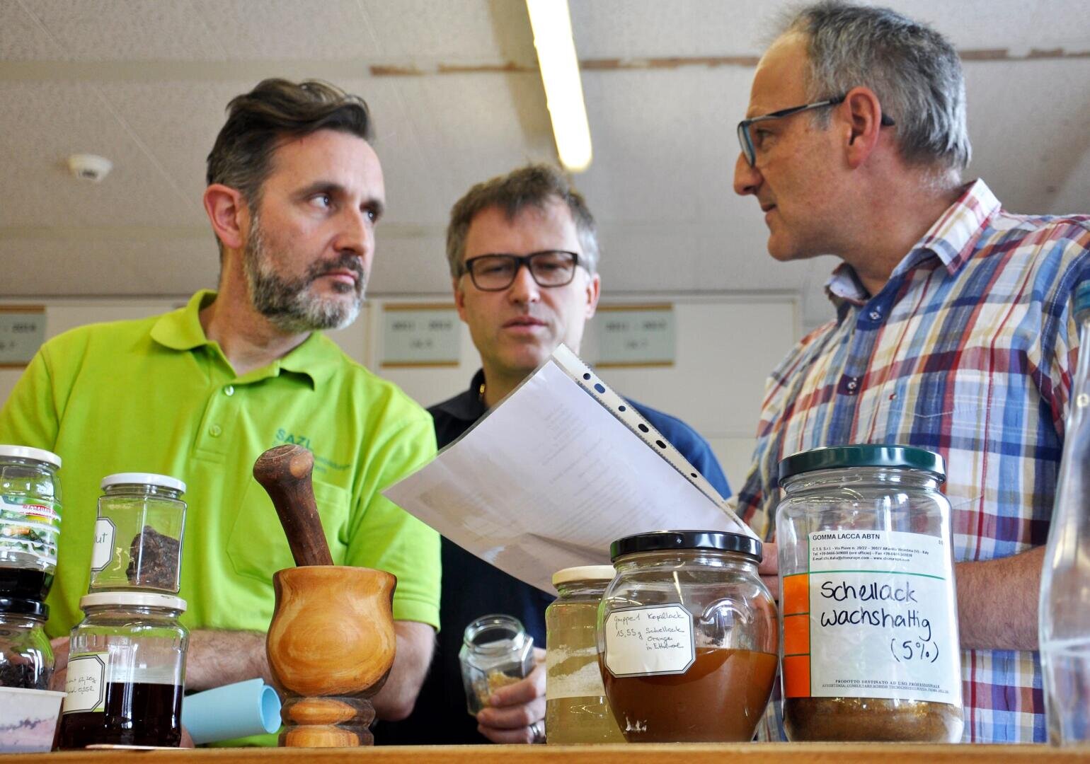 Drei Männer stehen hinter einem Tresen mit verschiedenen beschrifteten Gläsern und einem hölzernen Mörser, diskutieren etwas und halten Papiere in einer Labor- oder Werkstattumgebung.